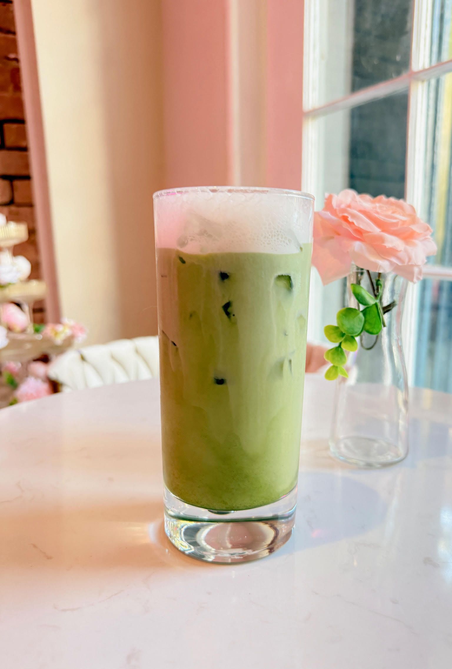 Iced matcha latte in a glass on a table, with a pink flower and window in the background.