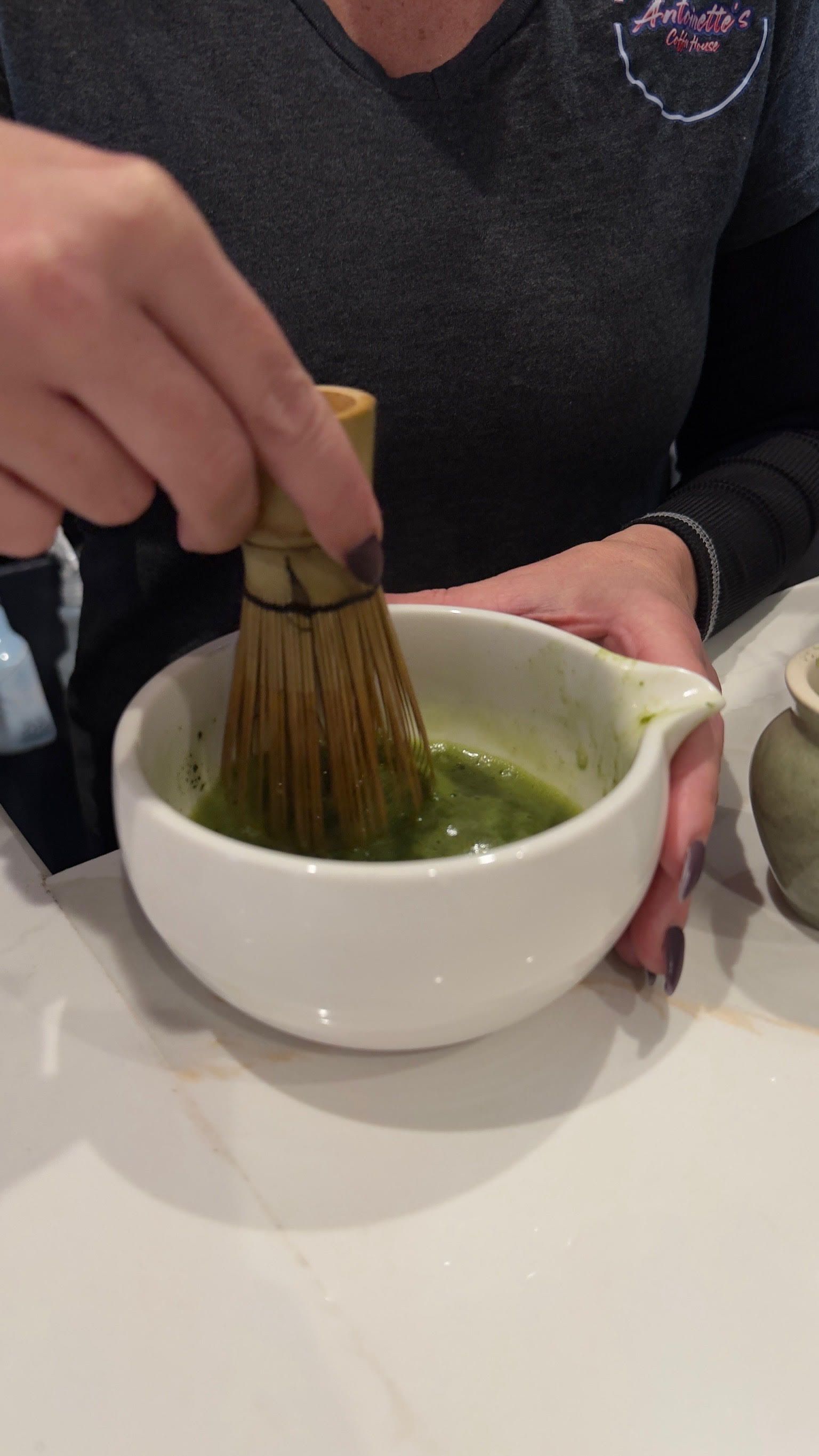 Person whisking matcha tea in a white bowl with a bamboo whisk. Green liquid.