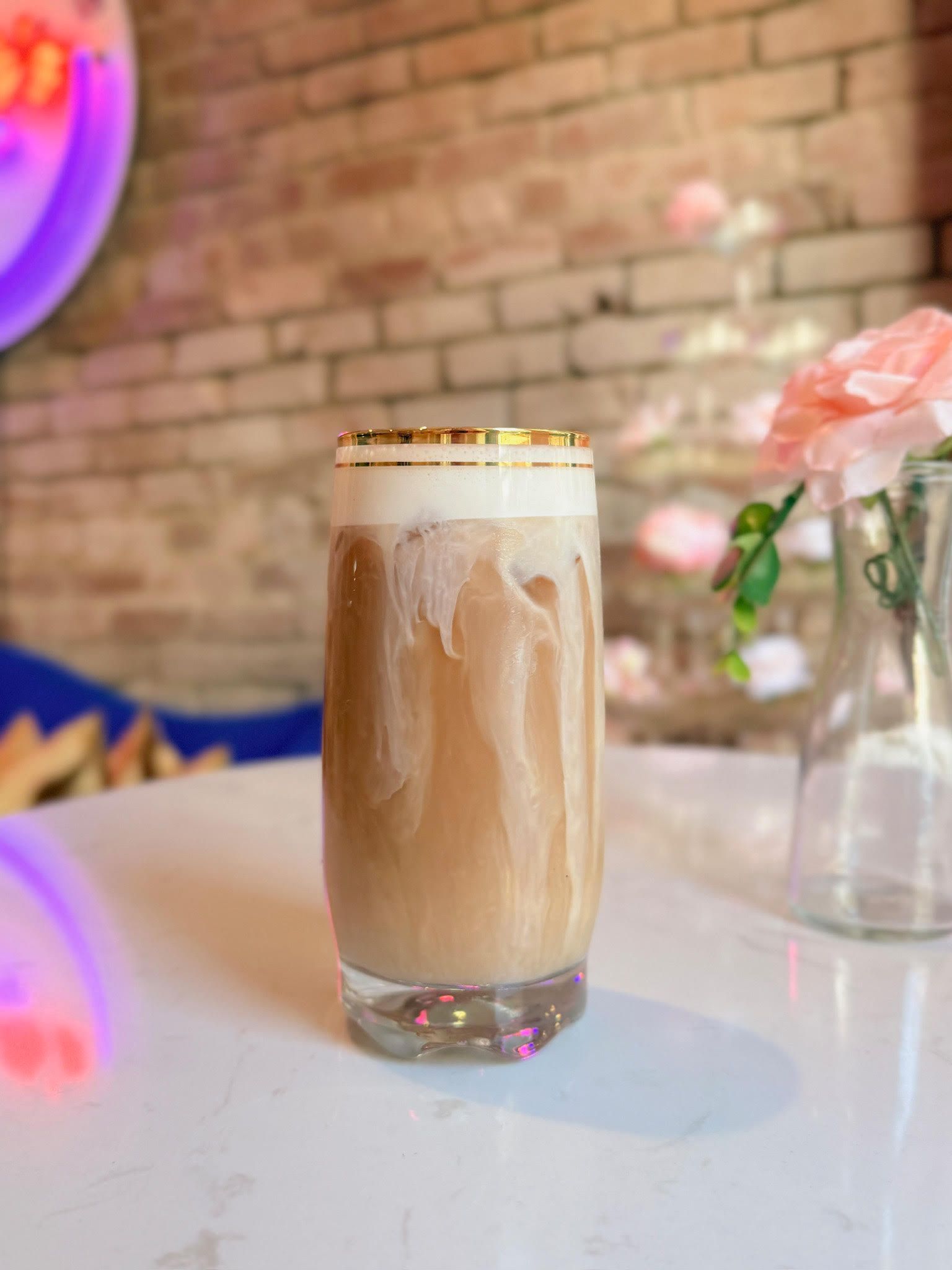 Iced coffee drink in a tall glass with cream, against a brick wall and flowers on a white table.