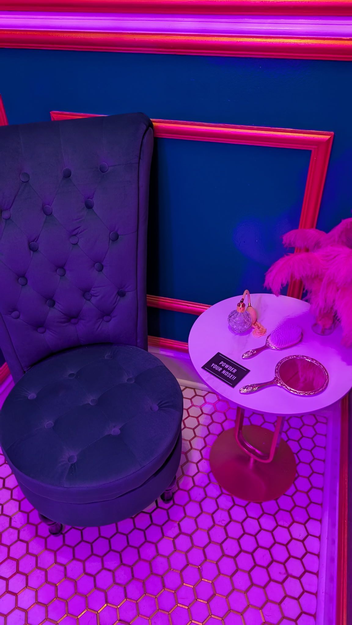 Purple-lit room with a high-backed tufted chair, small table with mirror, and decorative feather arrangement.