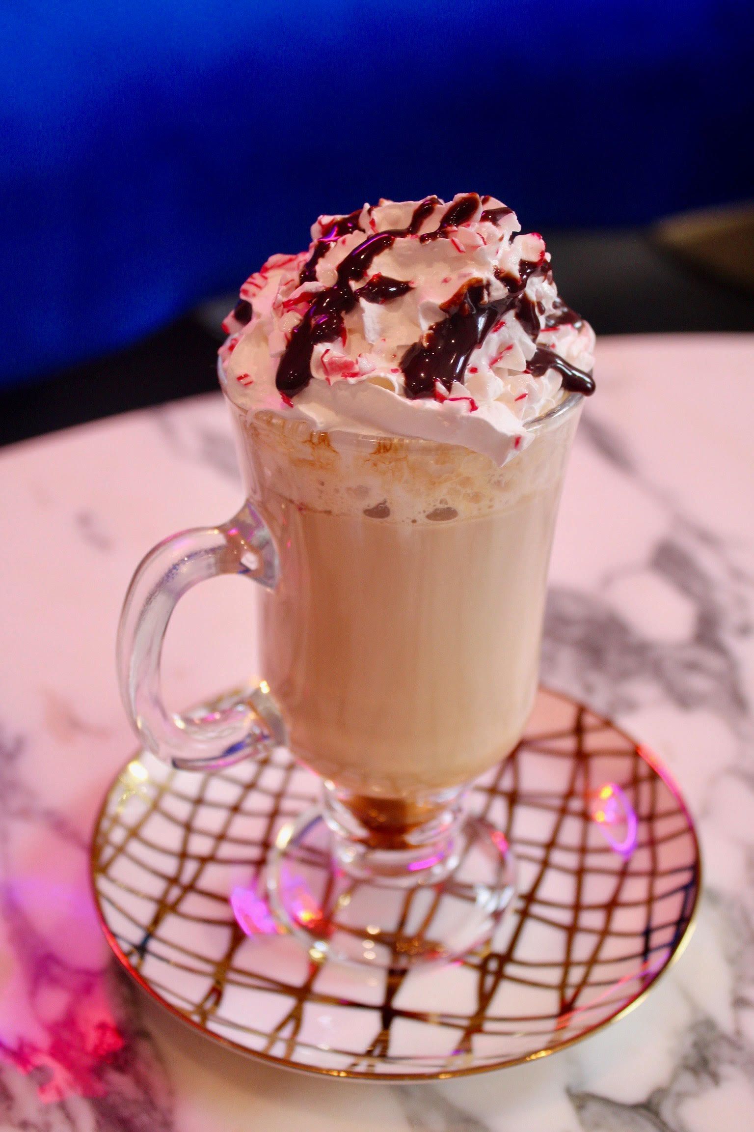 Coffee drink in clear glass, topped with whipped cream and chocolate drizzle, on patterned saucer.