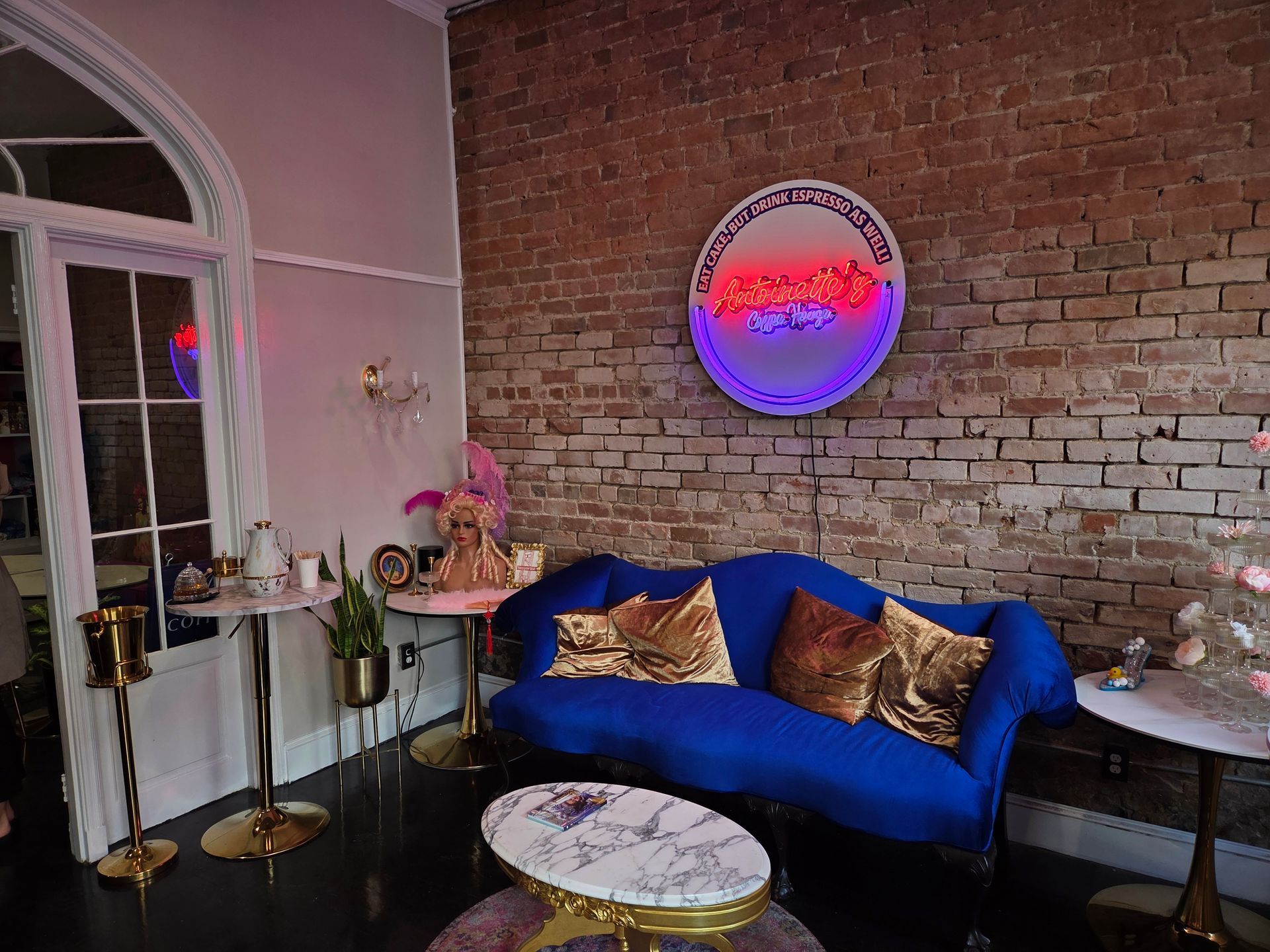 A blue velvet couch with gold pillows against a brick wall, with a neon sign.