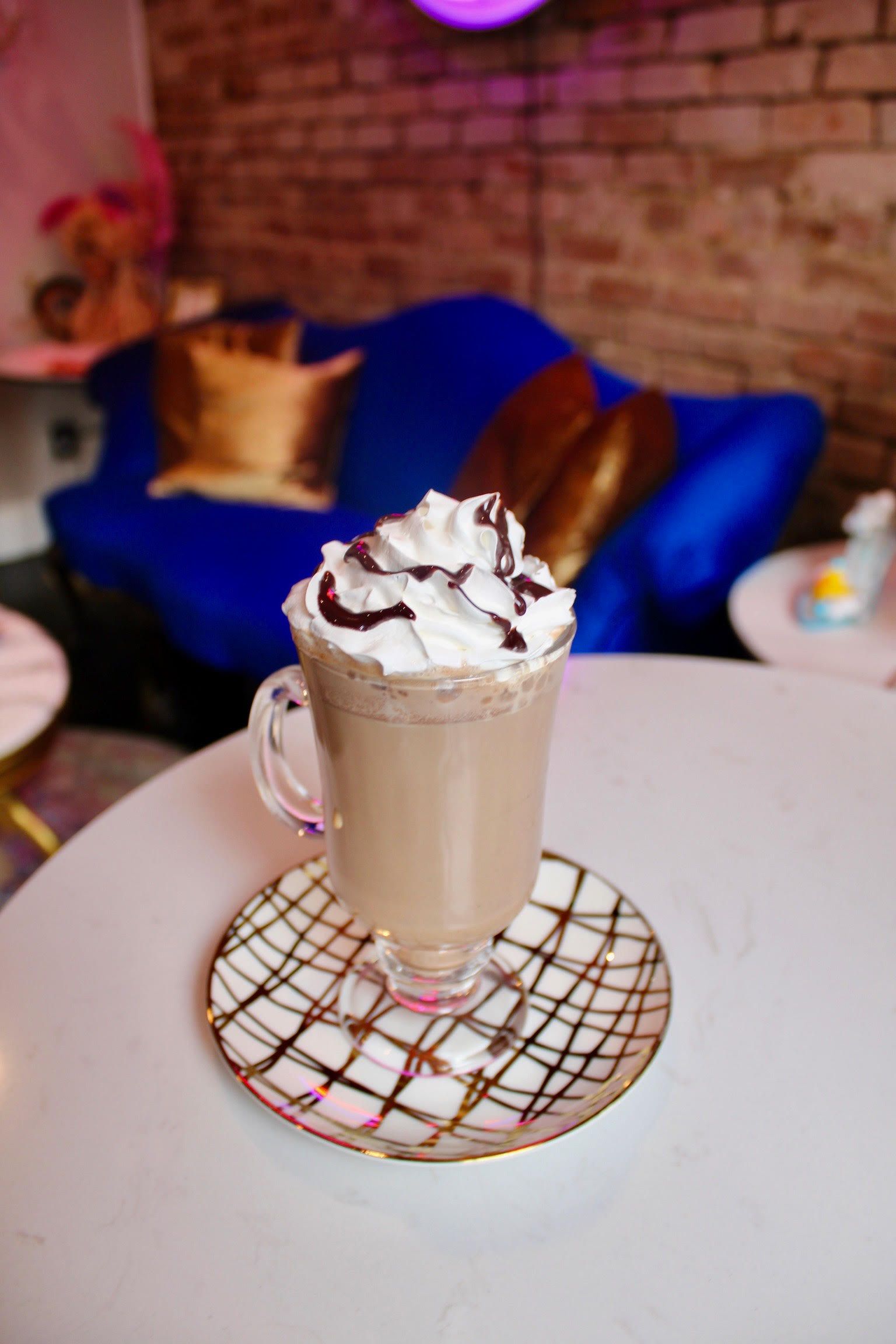 Iced coffee with whipped cream and chocolate syrup on a patterned saucer, sitting on a white table.