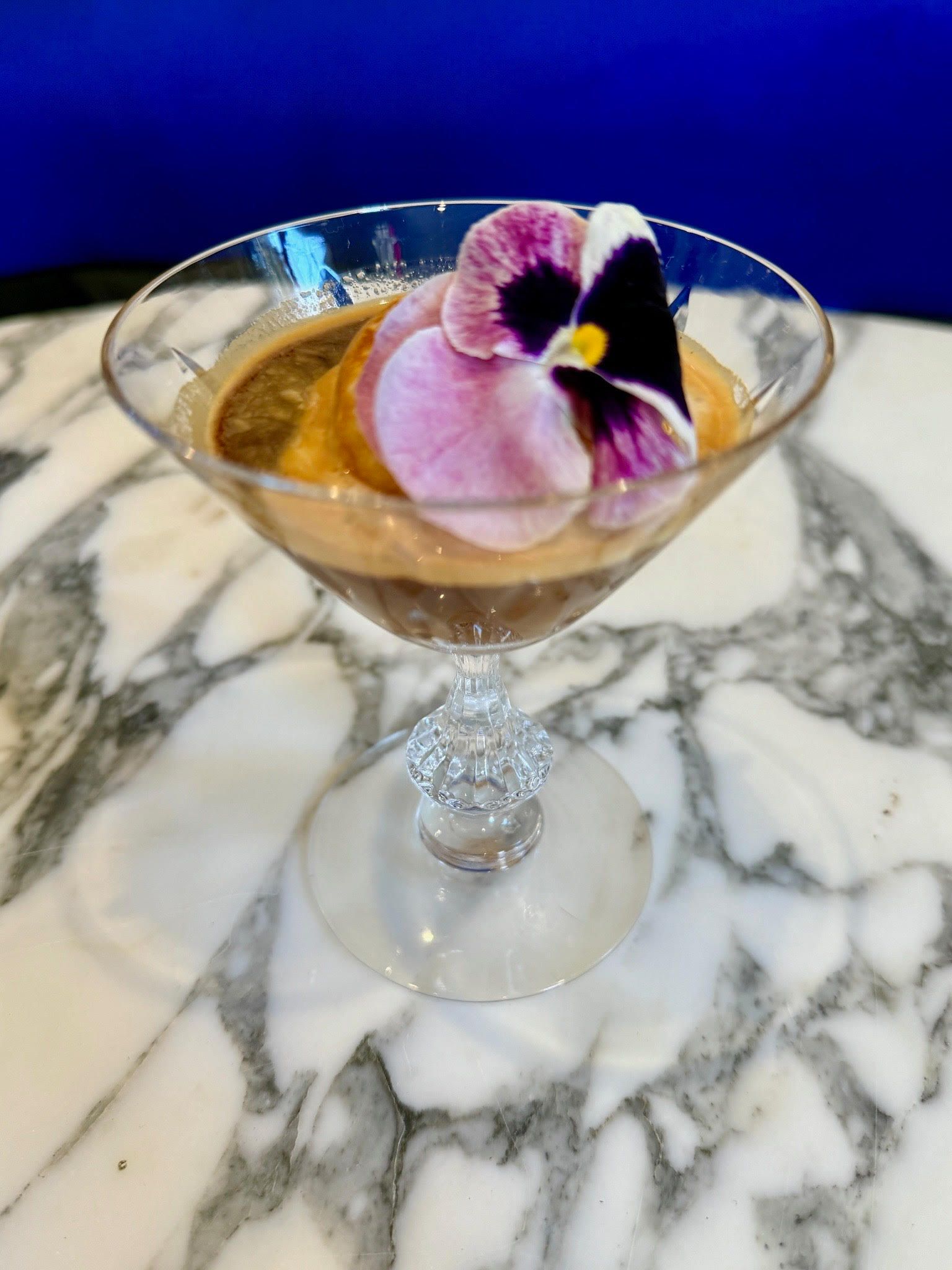 Dessert in a stemmed glass, topped with a purple and white flower, on a marble surface.