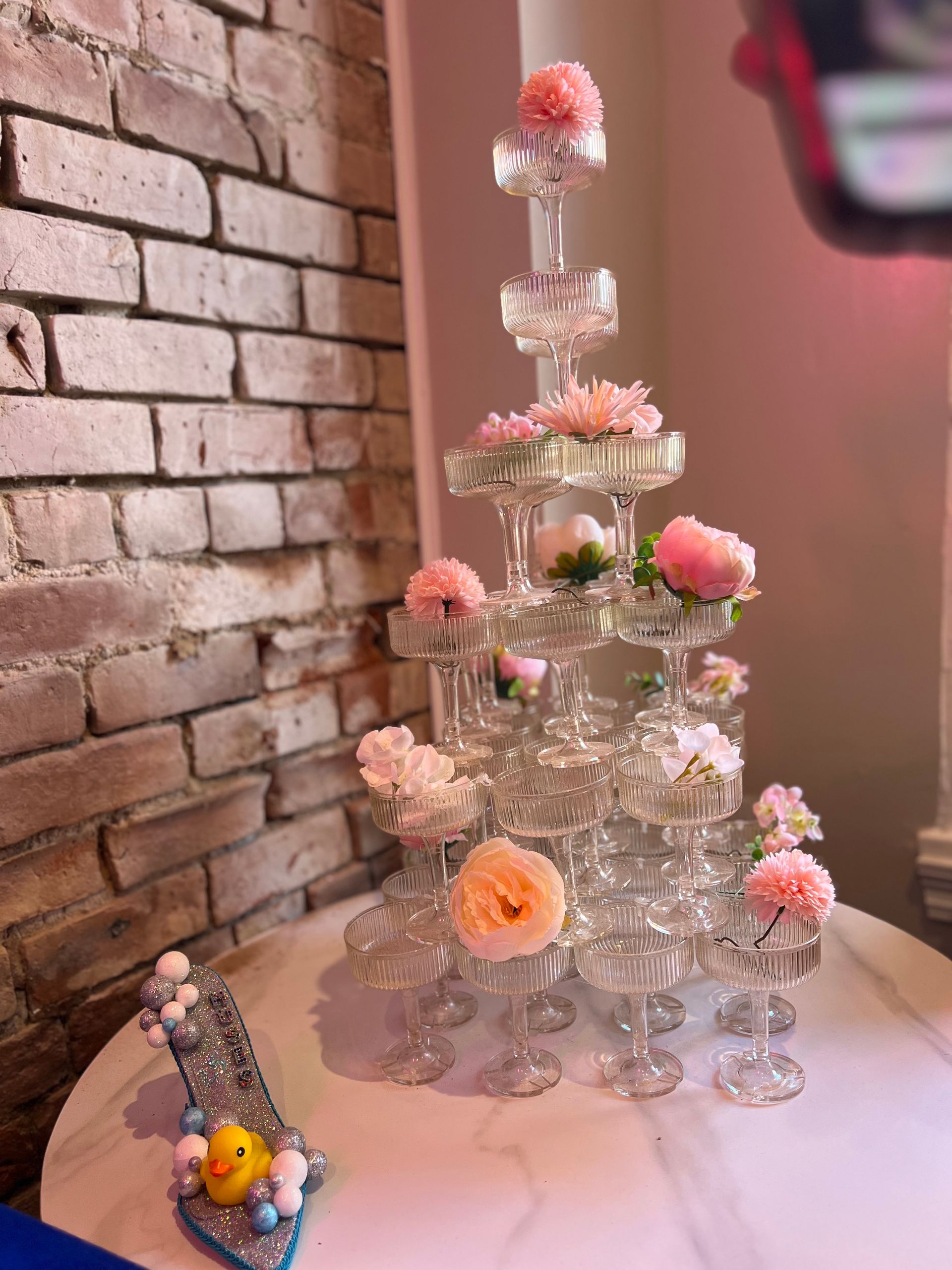 Champagne glass tower with pink flowers on a white table next to a brick wall. A sparkly shoe.