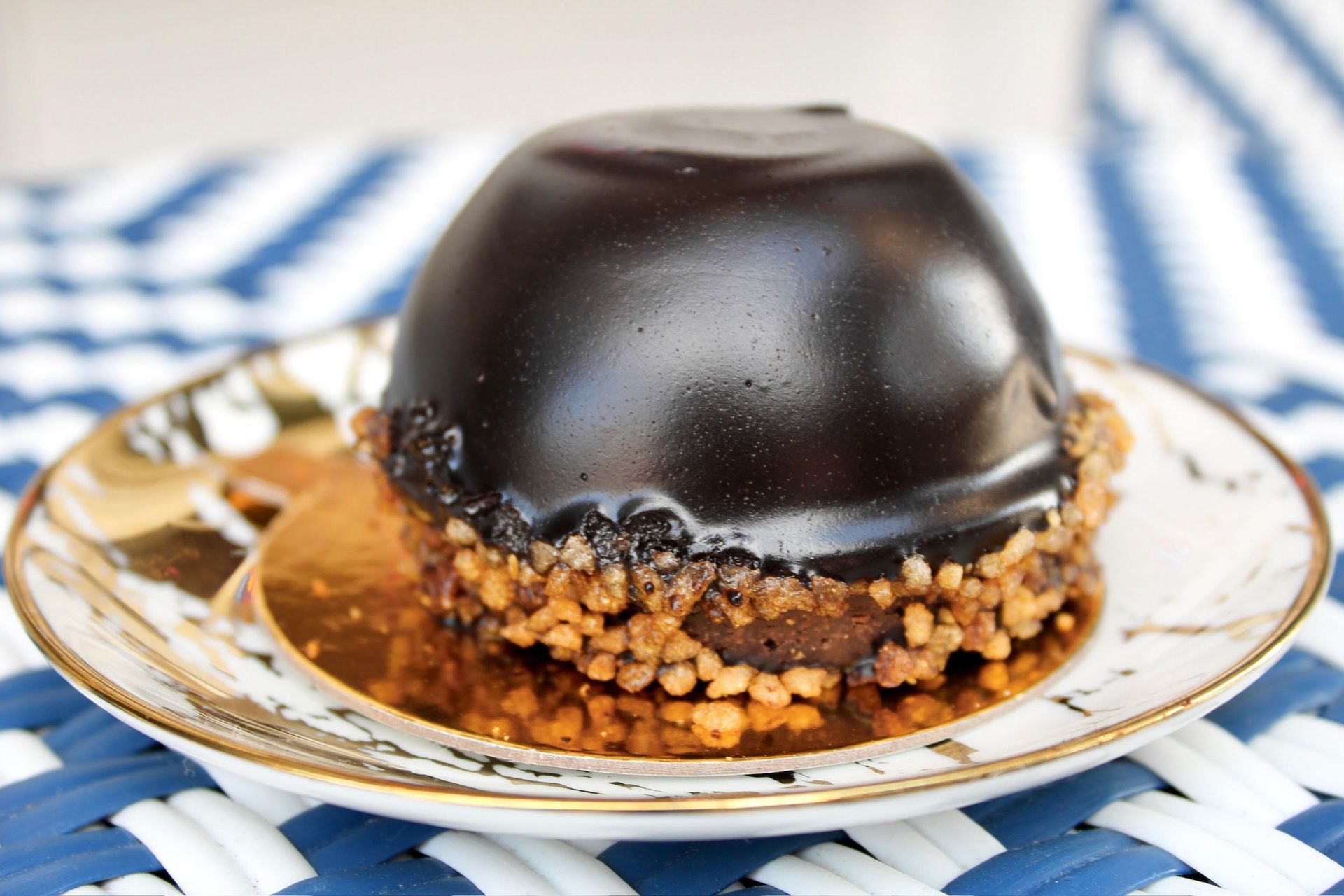 Chocolate dome pastry with nuts, on a gold-rimmed plate over a blue and white striped surface.
