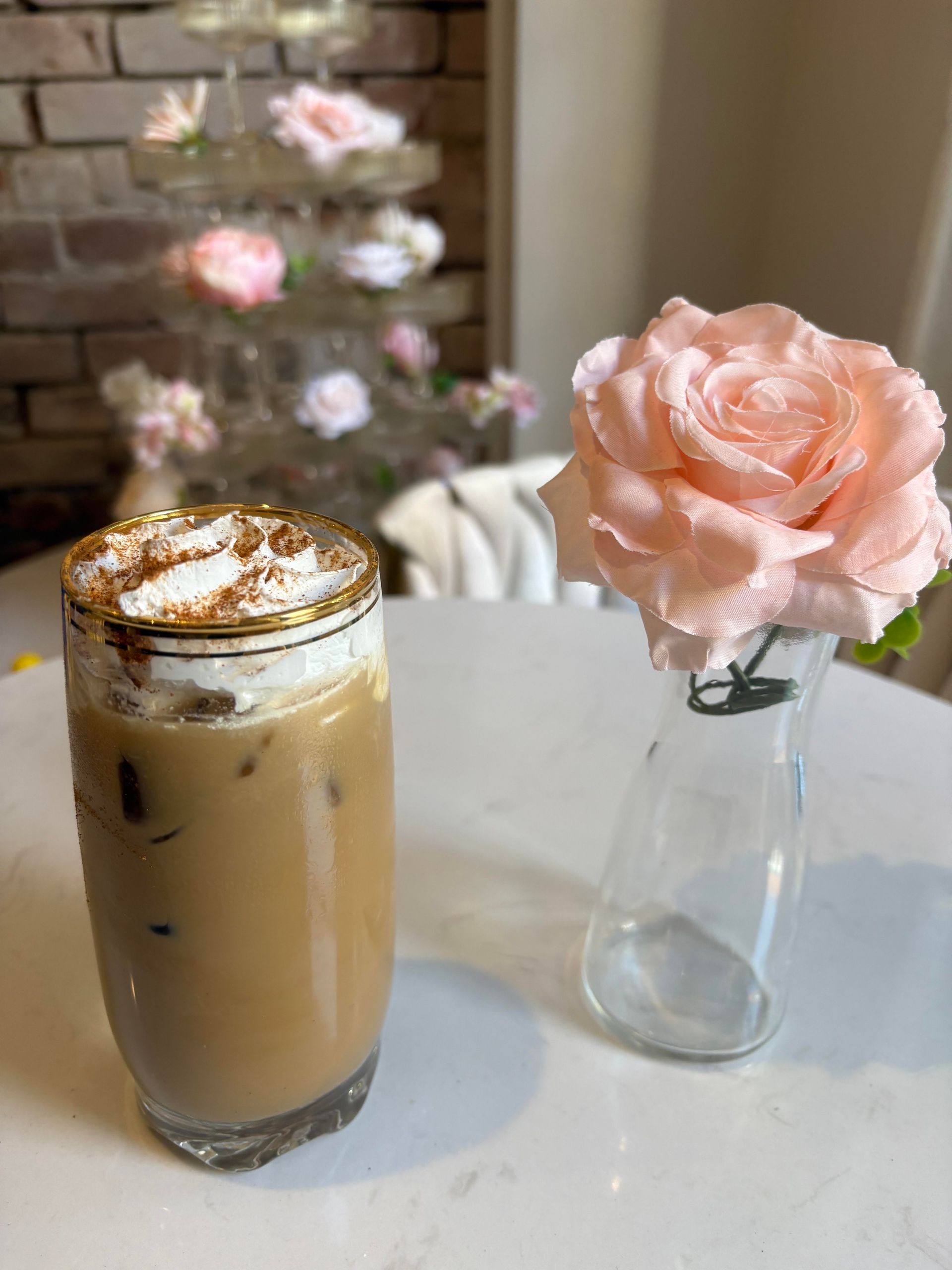 Iced coffee with whipped cream and a pink rose on a white table. A tiered floral display is in the background.