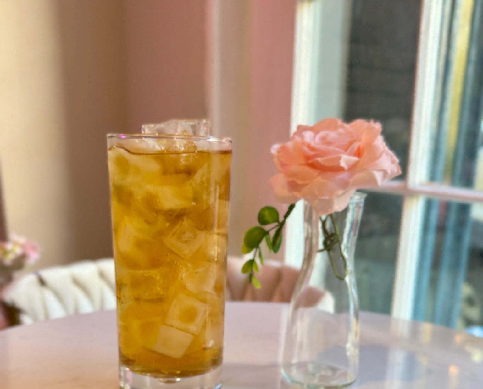 Tall glass of iced tea with ice cubes, next to a pink rose in a vase.