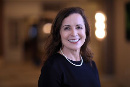 Woman with brown hair smiles, wearing a patterned blouse, standing in front of a gray wall.