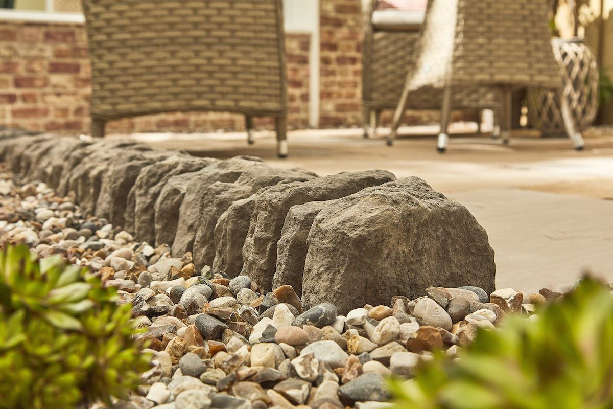 Stone border along a walkway, with gravel, plants, and wicker chairs visible in the background.