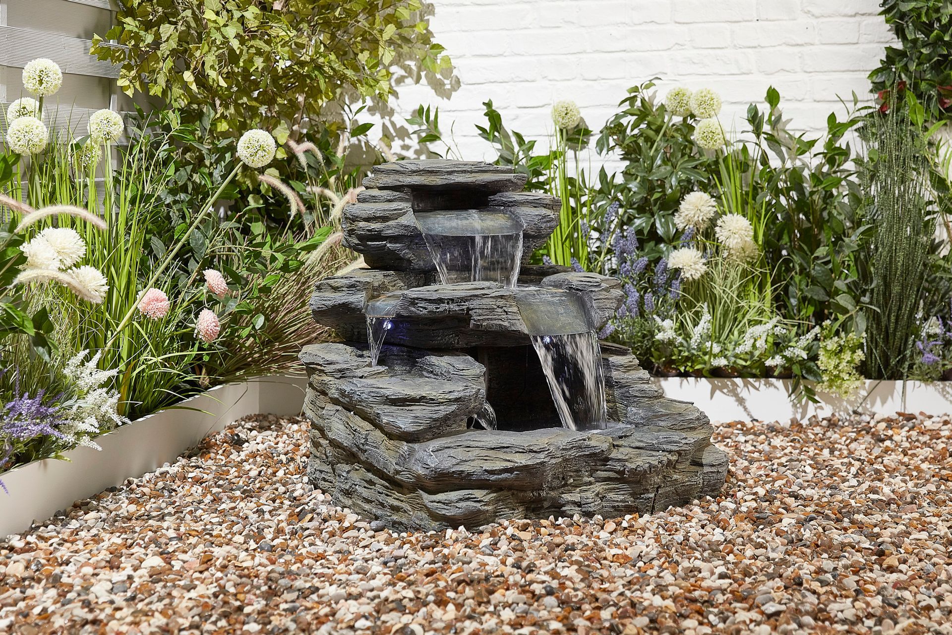Rock fountain in a garden setting, water cascading down levels into a basin, surrounded by plants and gravel.