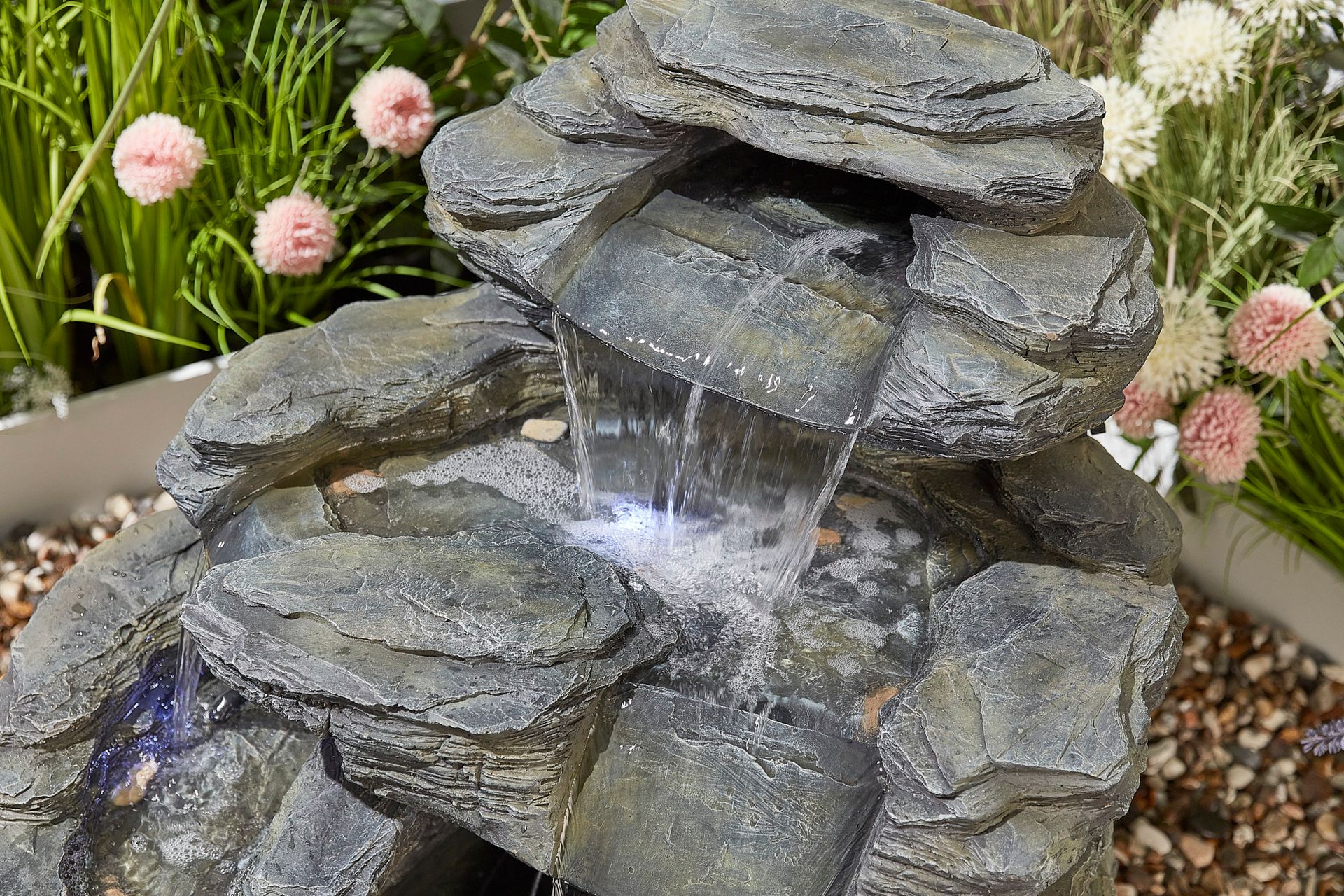 Rock fountain with cascading water, surrounded by green foliage and pink flowers.