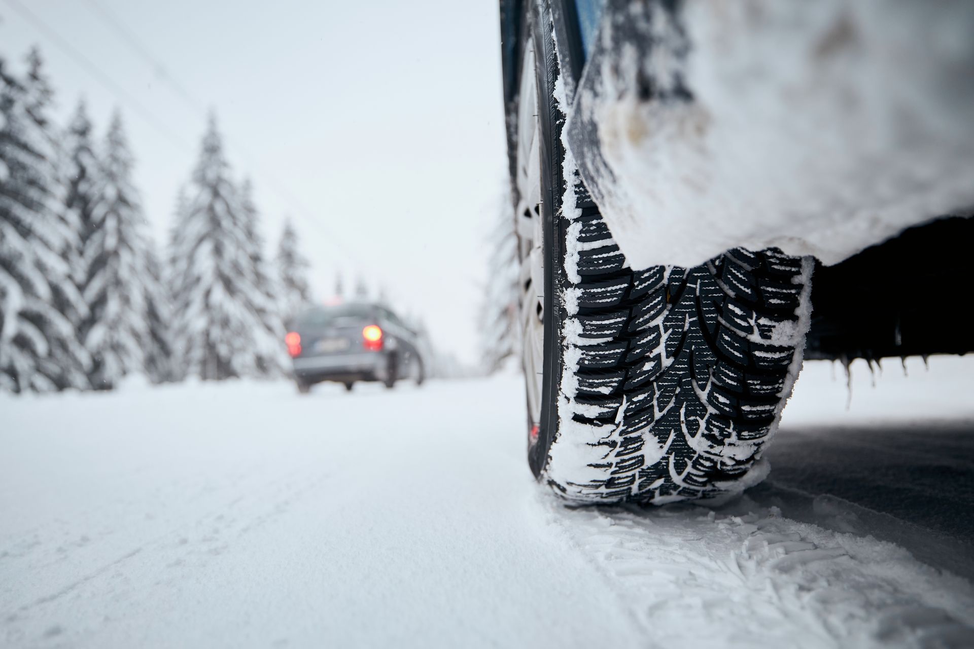 Close-up of a car tire on a snowy road, trees in background, another car further down the road.