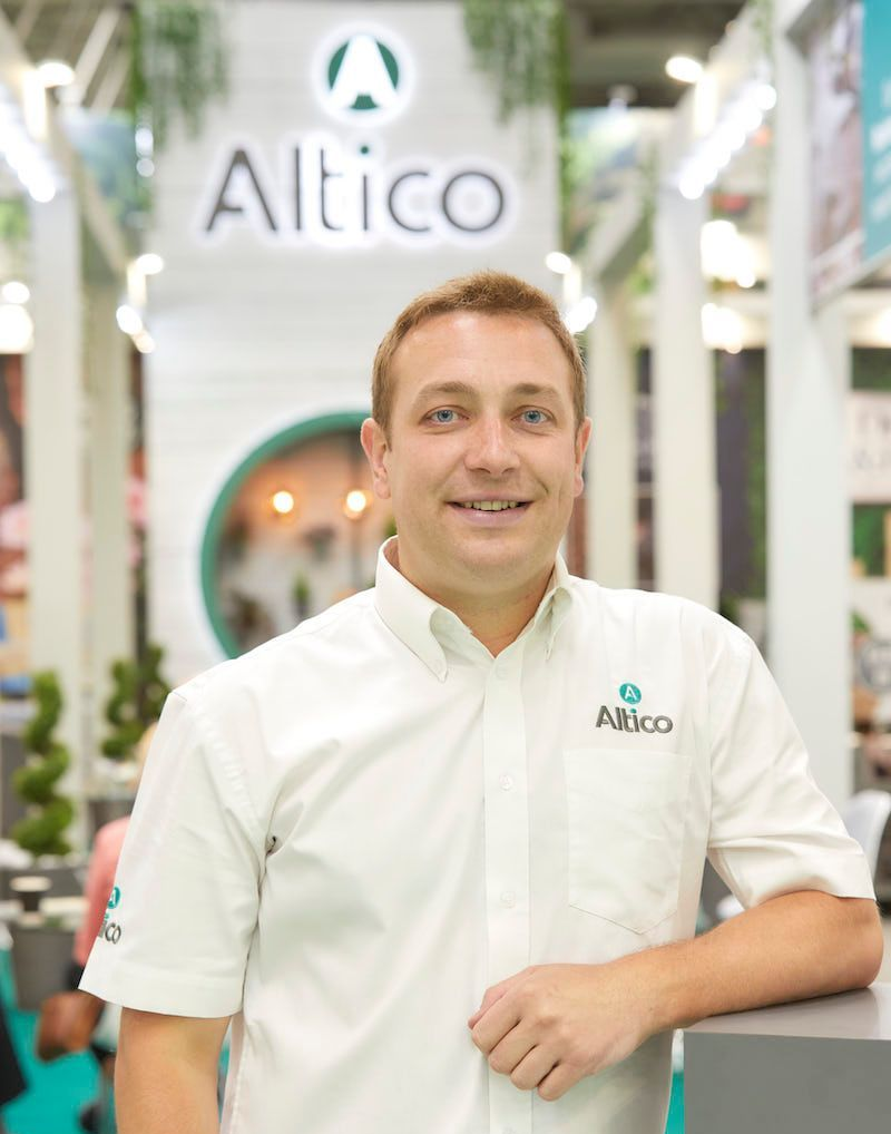 Man in white shirt with company logo, smiling. Leaning on gray counter, at a trade show with 