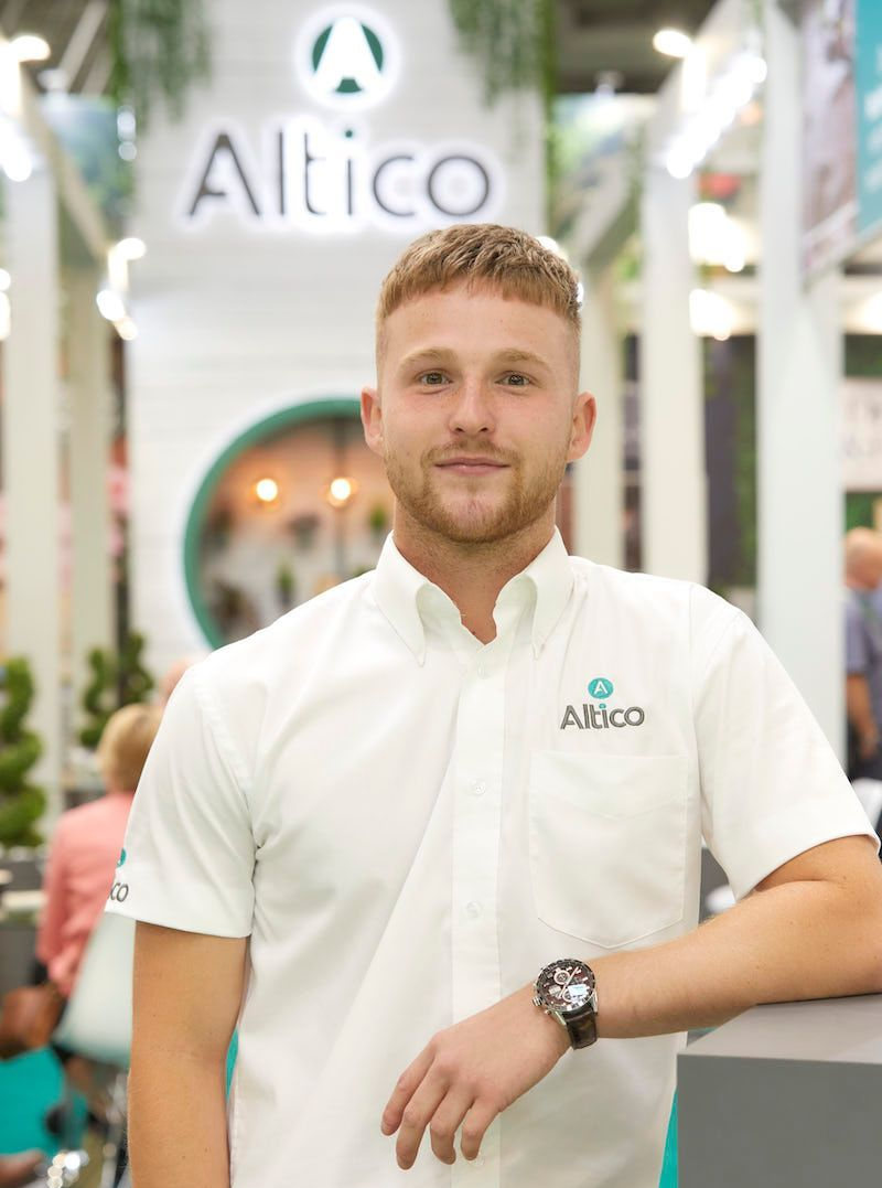 Man in white shirt with the Alitico logo. Stands in front of the Alitico sign at a booth, smiling.