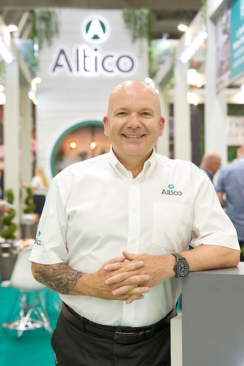 Man in white shirt with folded hands smiles at camera in front of the Altico logo at a trade show.