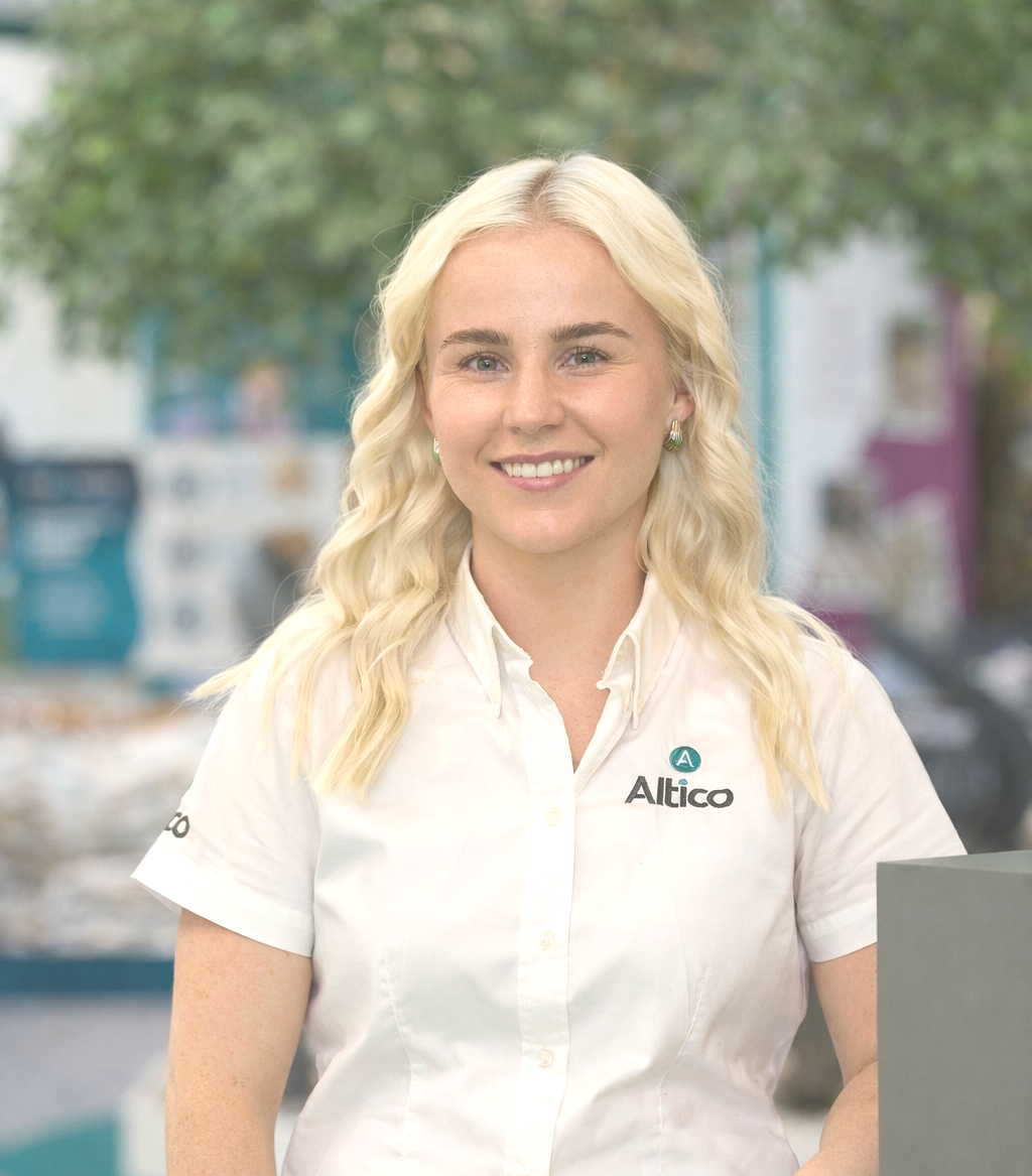 Blonde woman in white shirt and black skirt at an Altico trade show booth, smiling.