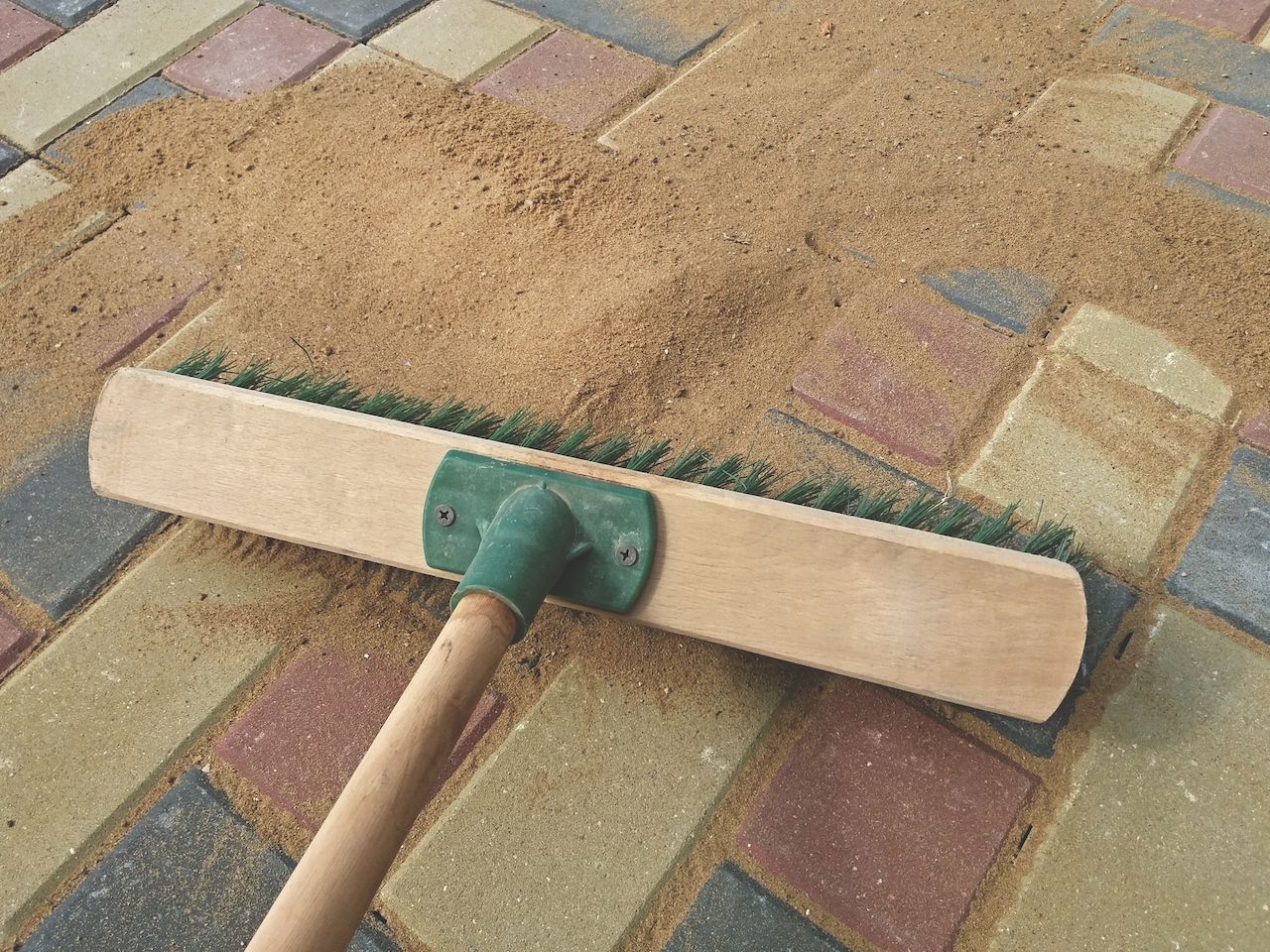Broom spreading sand over colorful paving stones, filling gaps in a walkway.