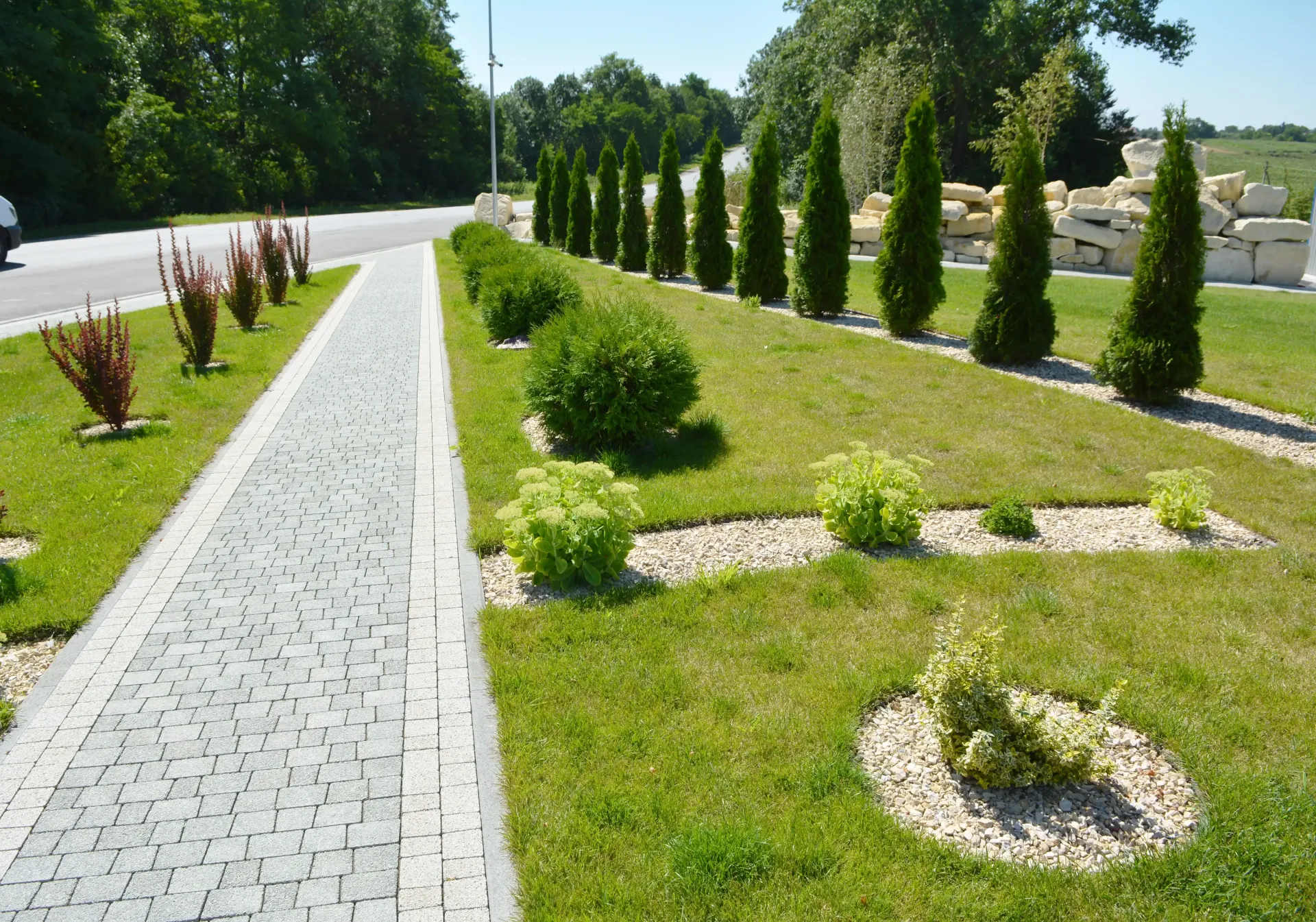 Curved stone walkway beside a manicured lawn with shrubs and tall trees on a sunny day