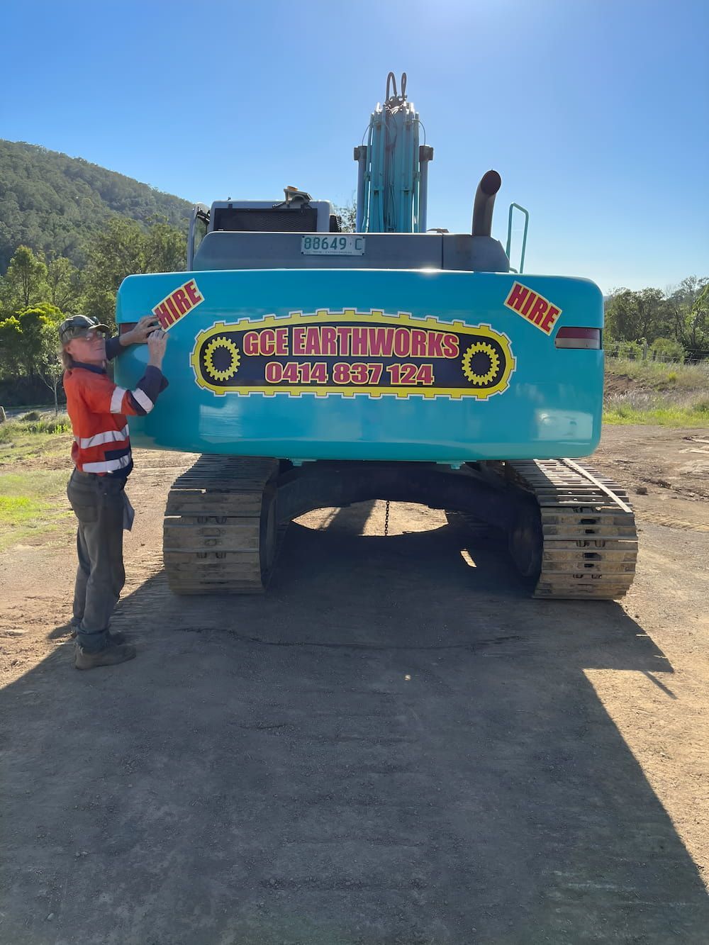 A Man Is Standing Next To A Large Blue Excavator That Says Hire On It — GCE Earthworks Pty Ltd In Krambach, NSW