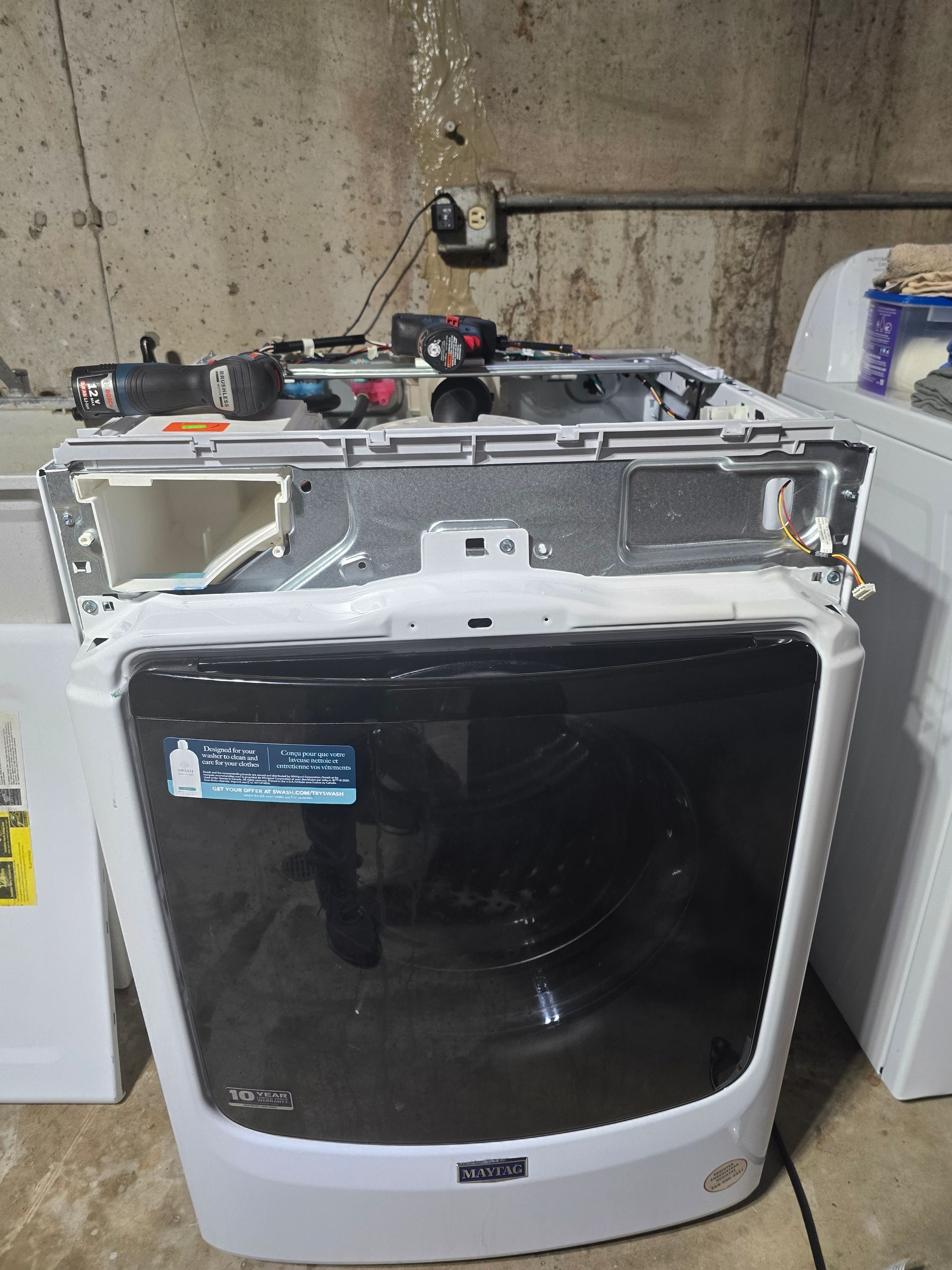 Maytag washing machine with the top panel removed, revealing internal components. In a concrete-walled room.