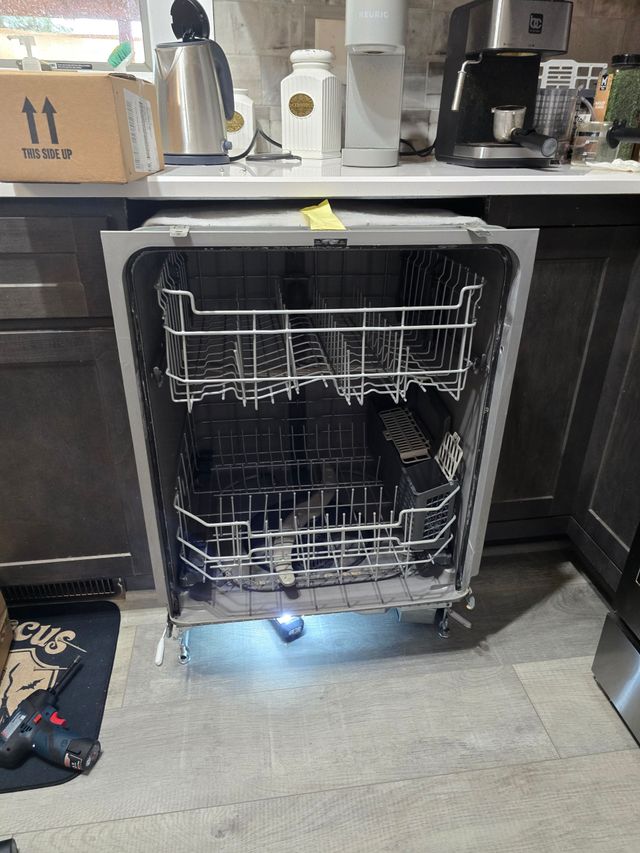 Open dishwasher with racks, a gray interior, in a kitchen with a countertop and cabinets.