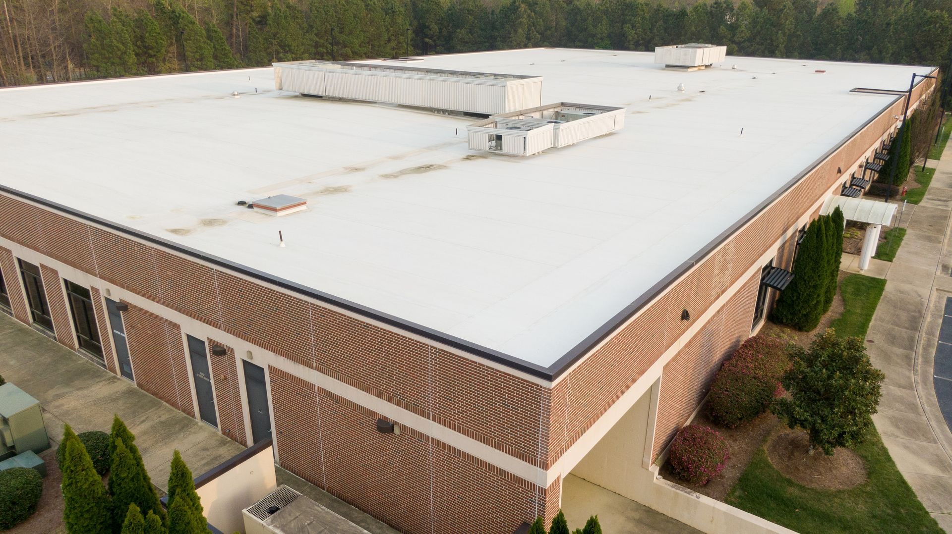 Aerial view of a brick industrial building with a white flat roof and surrounding trees