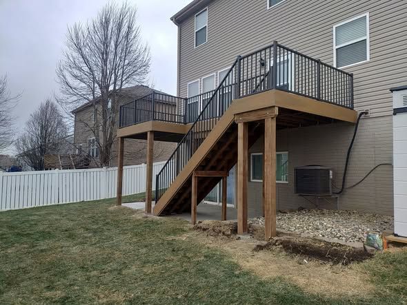 Two-story wooden deck with black railing and stairs attached to a beige house, surrounded by a yard with a white fence.