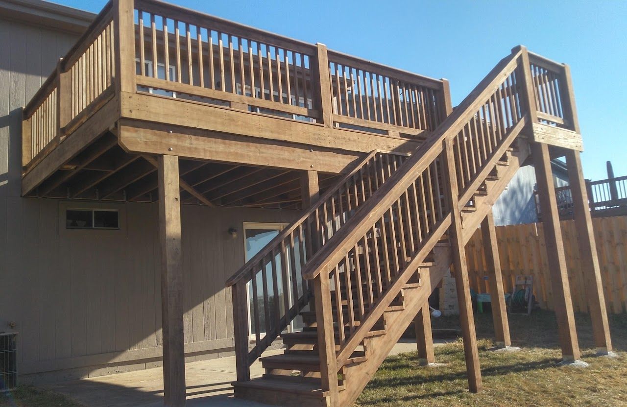 Wooden deck with stairs attached to a tan-colored house. Brown stained wood on a sunny day.