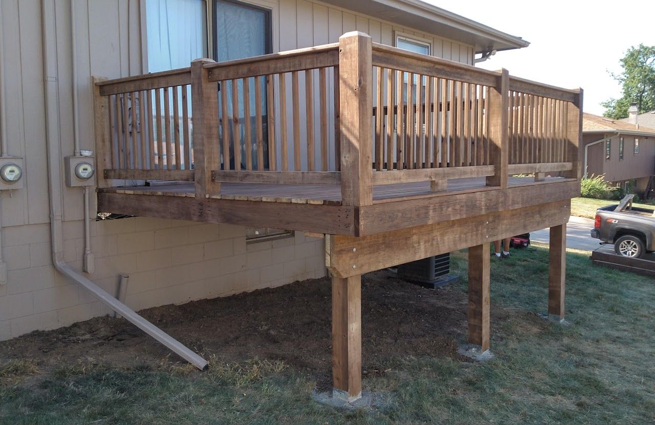 Wooden deck attached to a building, supported by posts. The deck is brown, in a grassy backyard.
