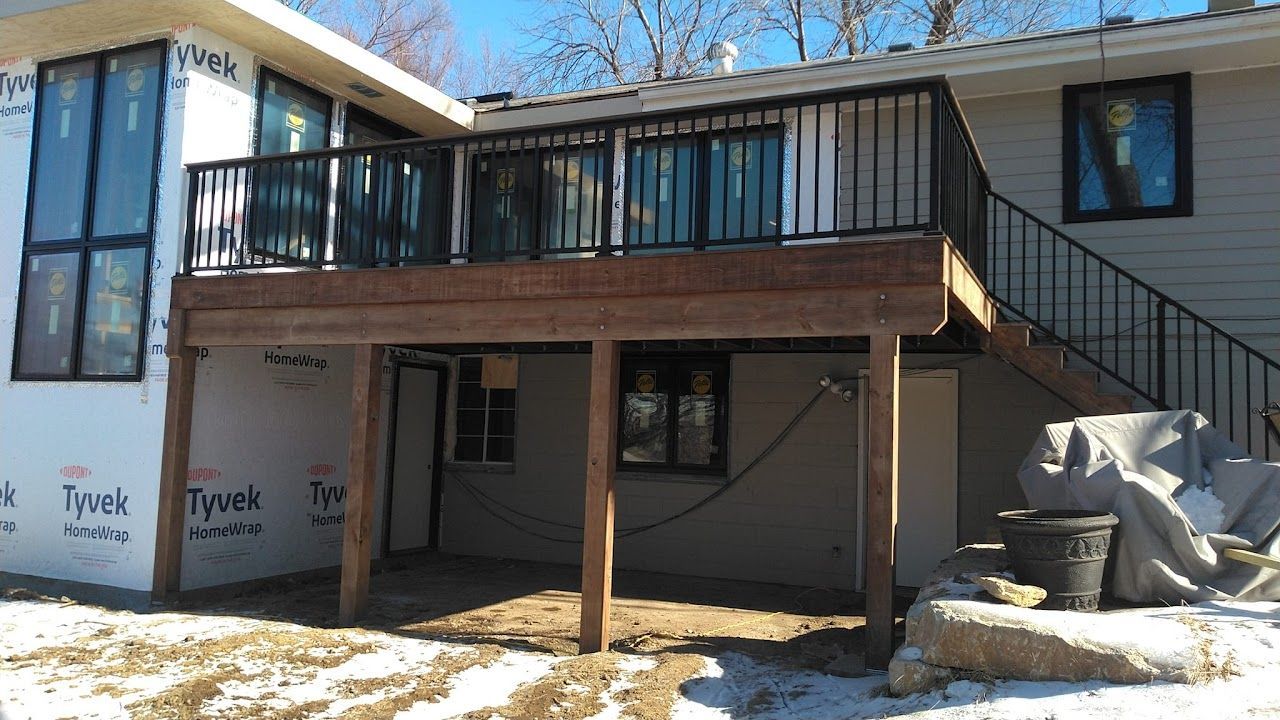 Deck with black railing, overlooking a shaded area. Wooden supports, black windows, and a black exterior staircase.