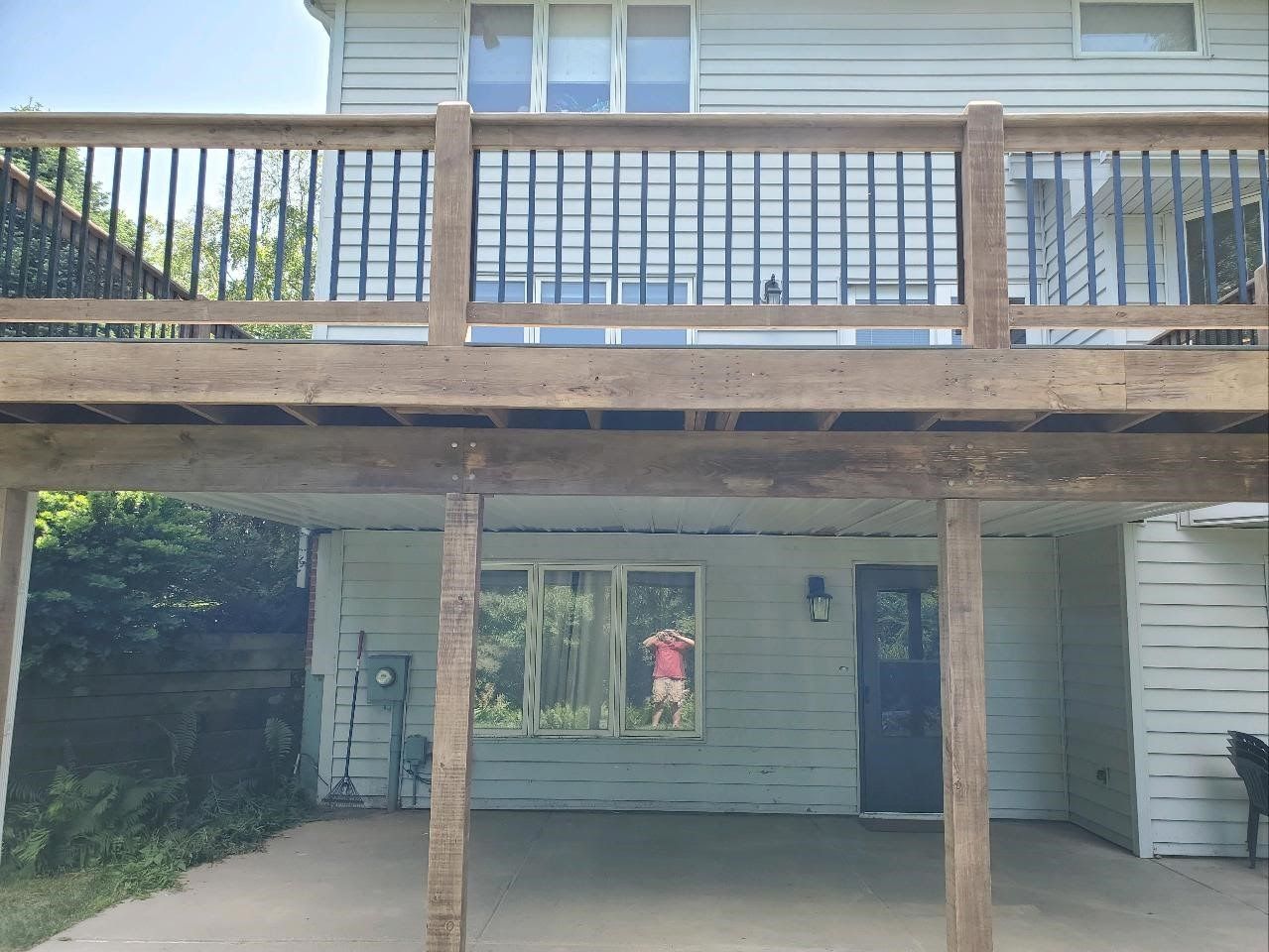 Two-story home exterior with a deck above a covered patio. Deck has brown railing, below is a concrete patio and window.