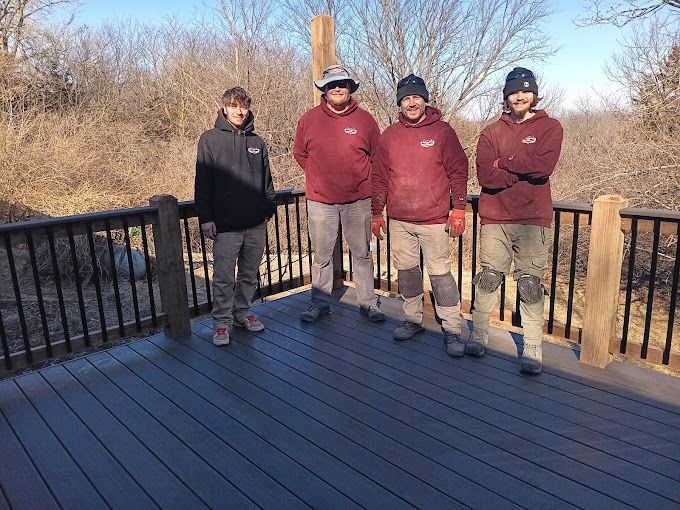 Wooden deck with pergola, dark railing, overlooking a backyard with a white fence.