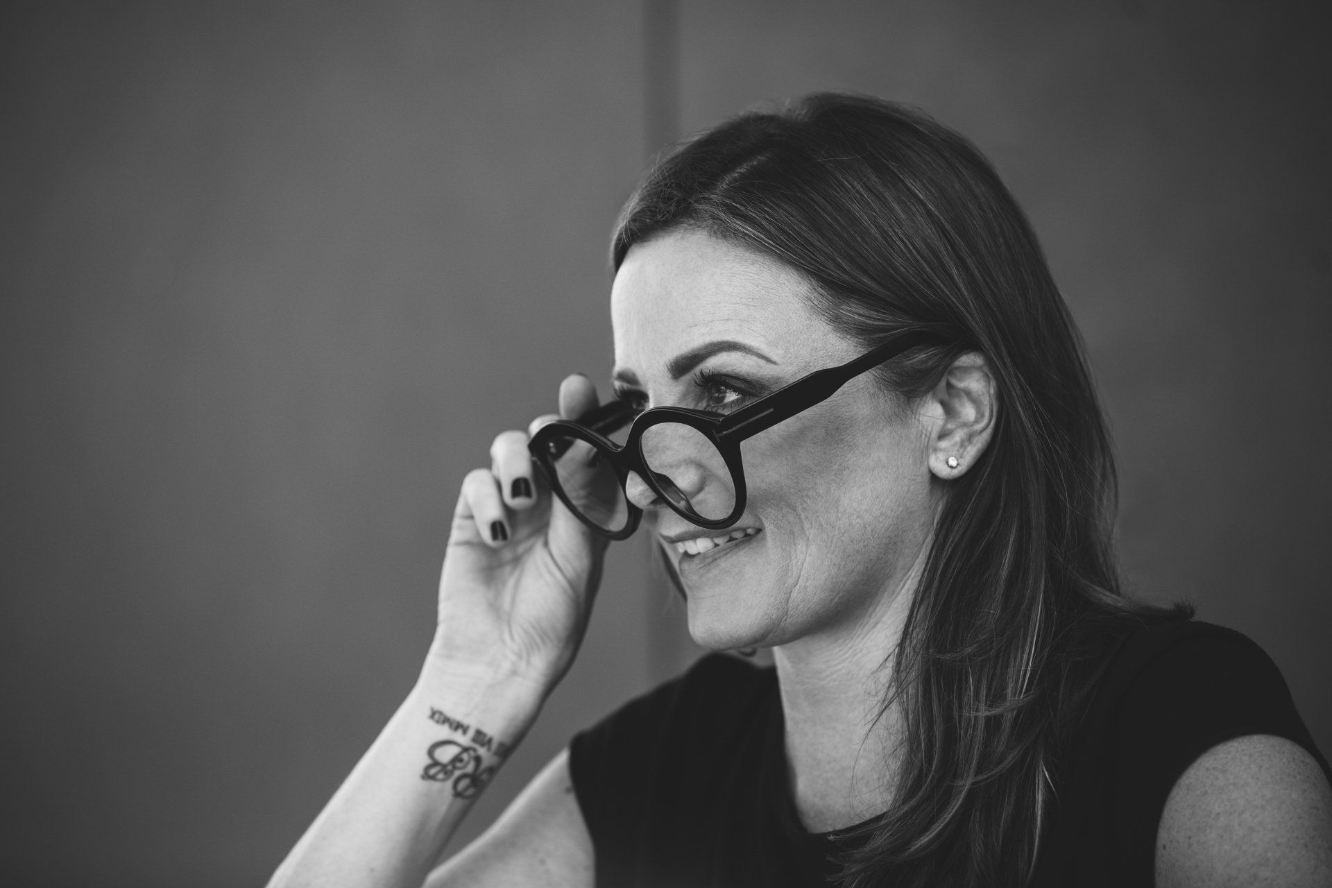 Eine Frau mit Brille und einer Tätowierung am Handgelenk lächelt auf einem Schwarzweißfoto.