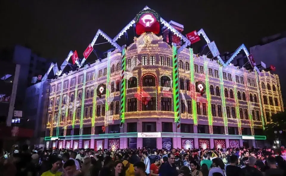 Uma multidão de pessoas está reunida em frente a um prédio que fica iluminado à noite.