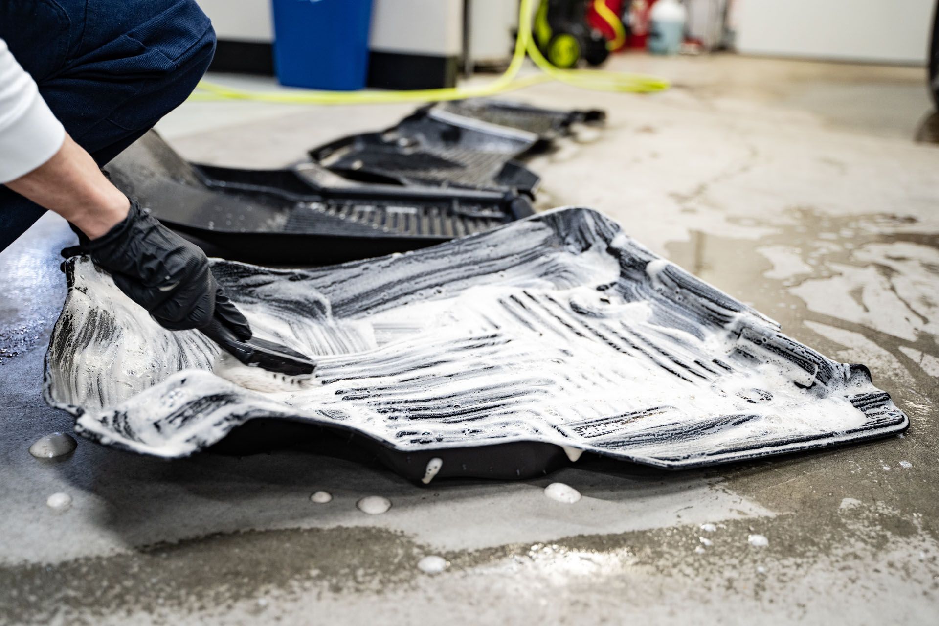 Person scrubbing black car floor mats with a brush and soapy foam in a garage.