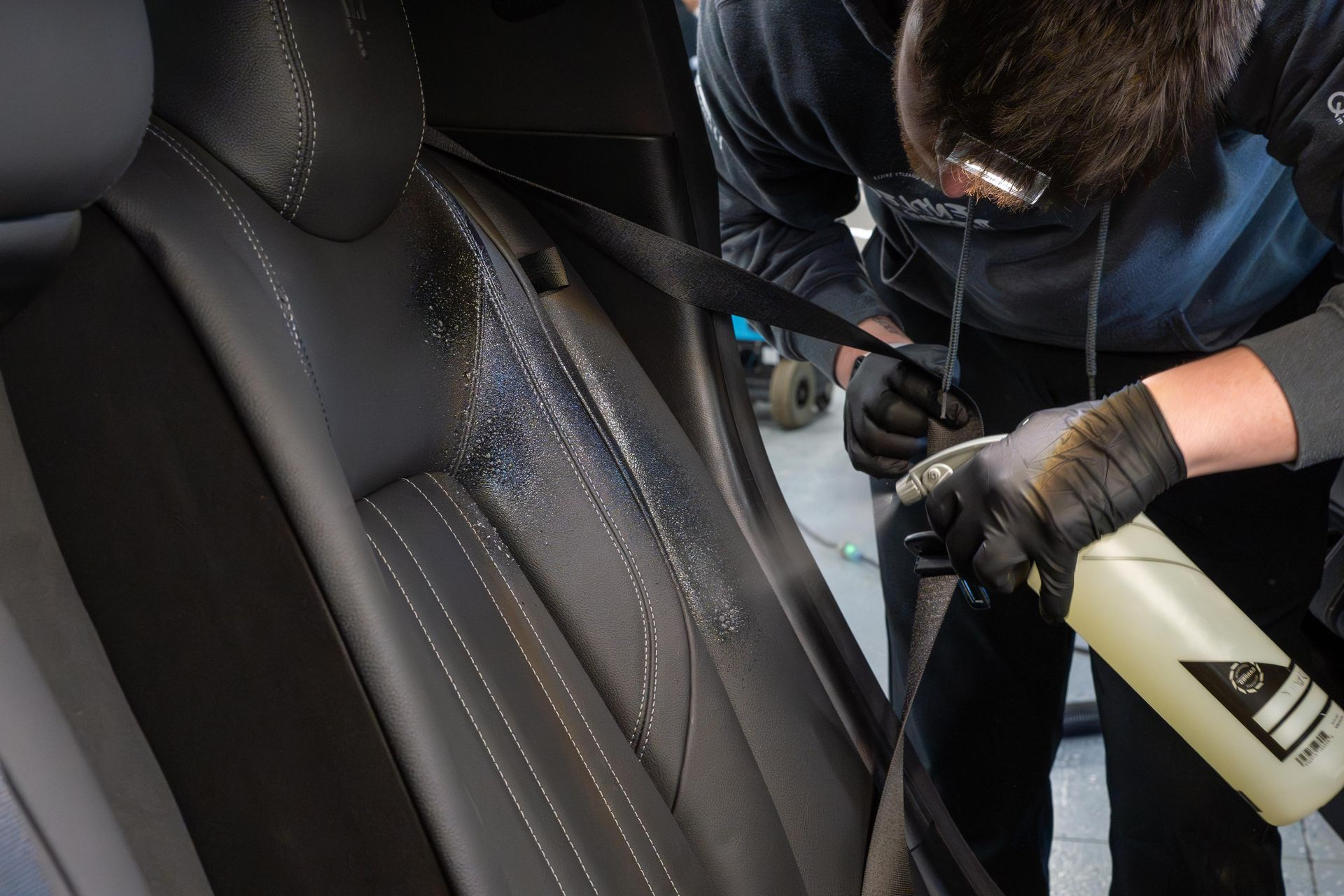 A person cleaning a car interior with a blue cloth and spray bottle, wearing black gloves.