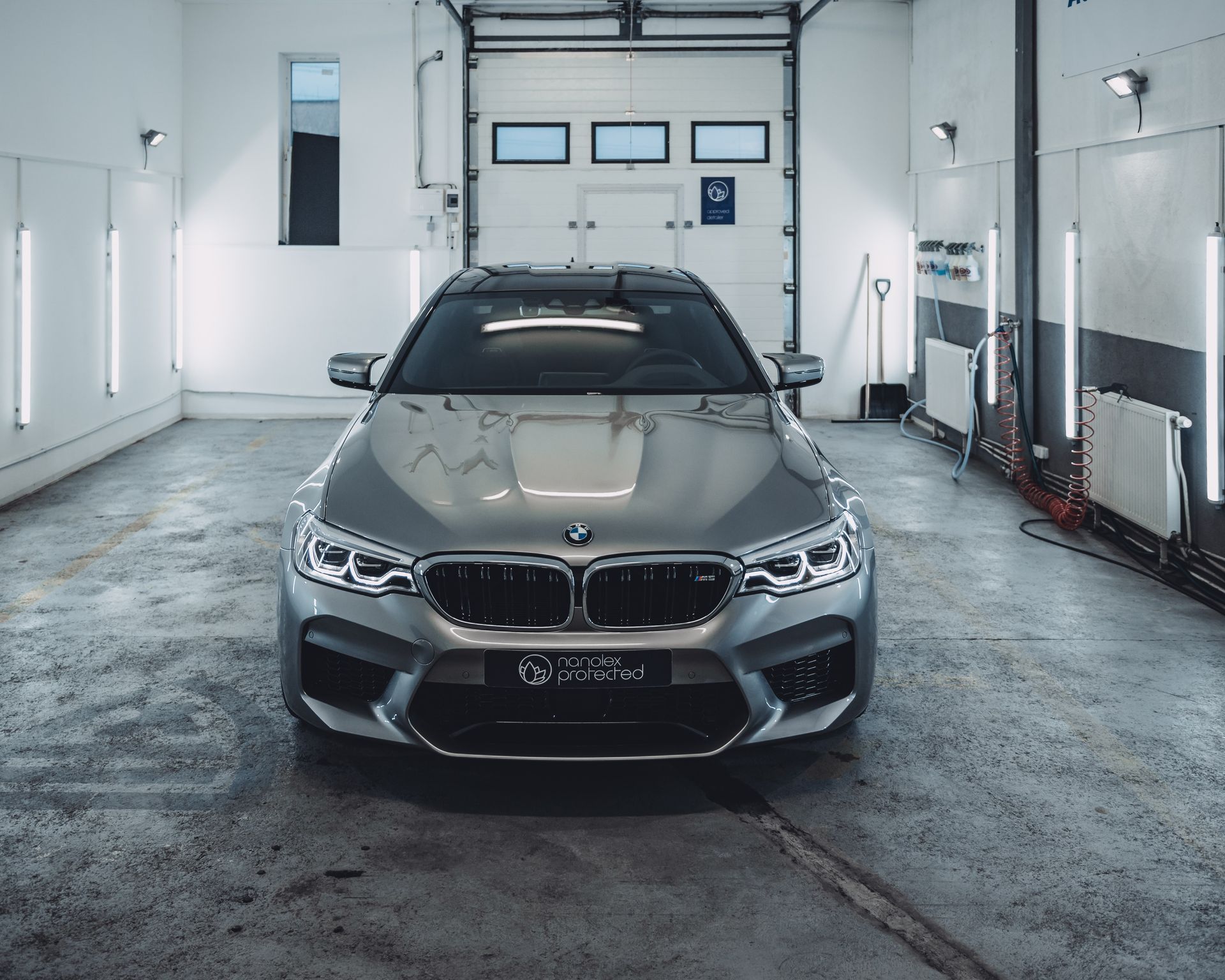 Silver BMW M5 in a garage with bright lights, centered in front of the closed door.