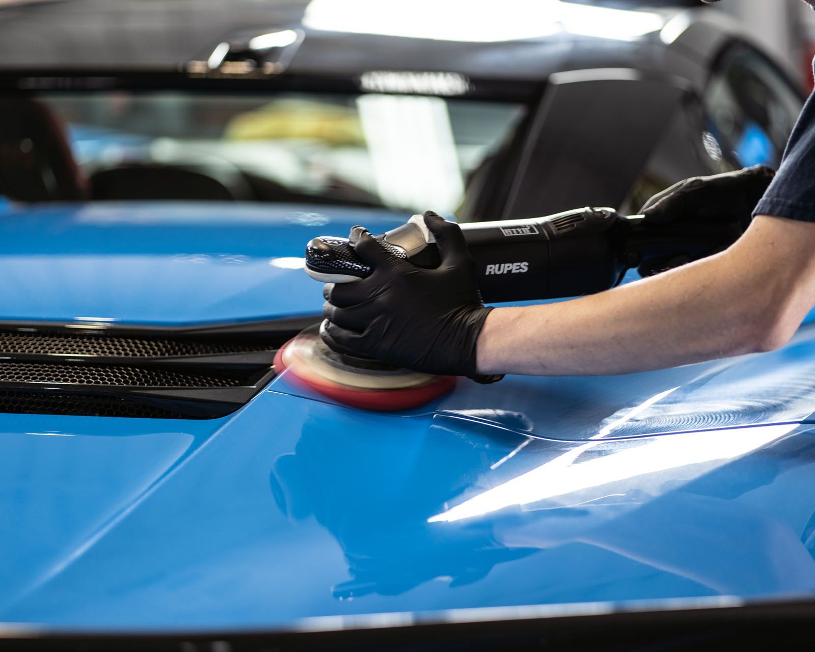 Person polishing a blue car with a machine buffer, wearing black gloves.