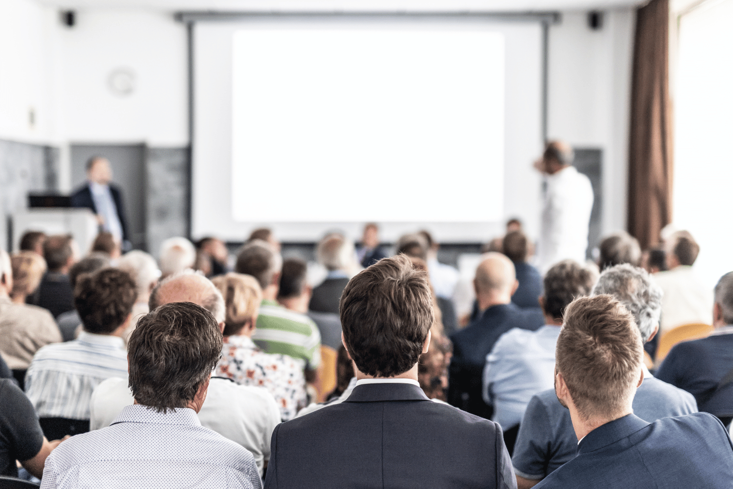 A rear view of an audience sitting in a seminar room, facing a large, blank projection screen and a speaker.