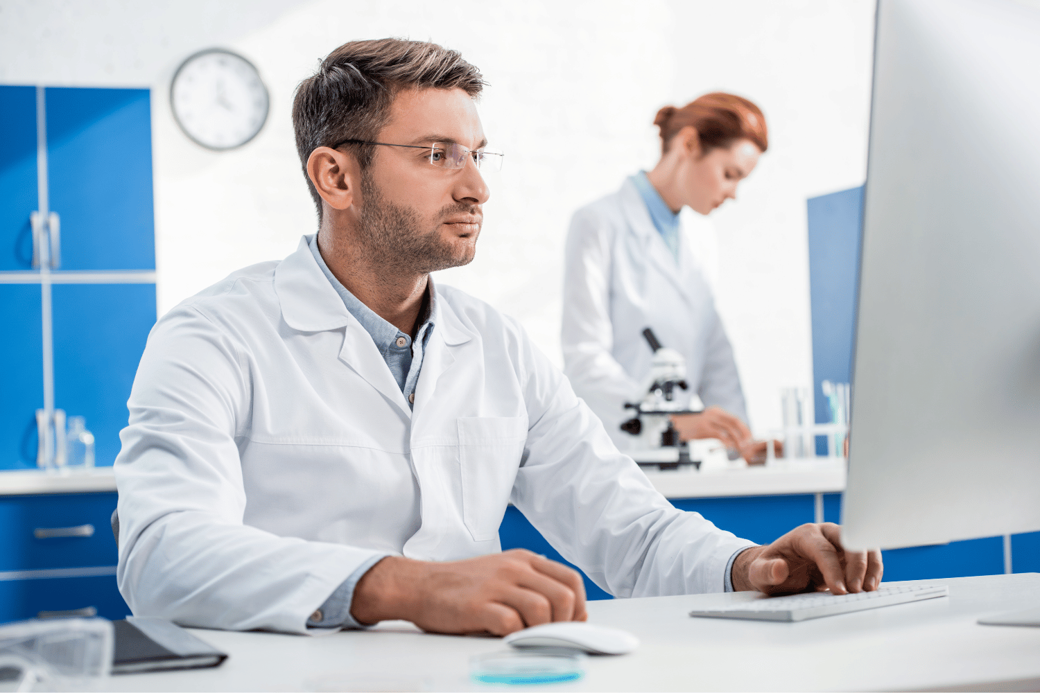 Two scientists in white lab coats work in a laboratory, with one focusing on a computer screen and another using a microscope.