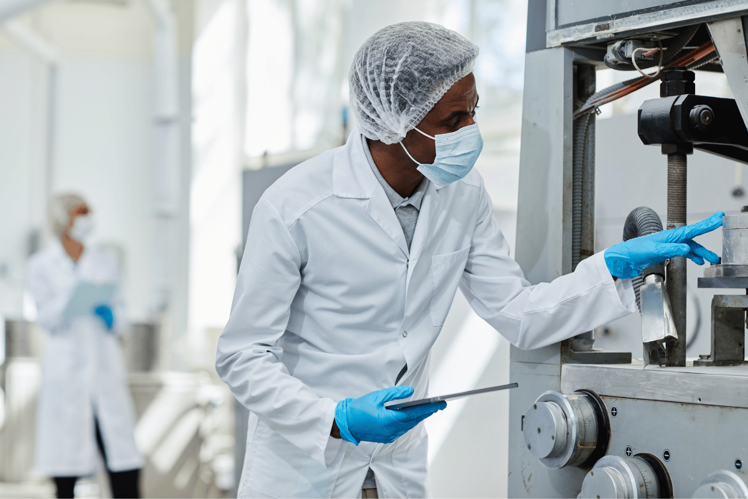 A scientist in a lab coat, mask, and hairnet inspects industrial machinery while holding a tablet in a laboratory setting.