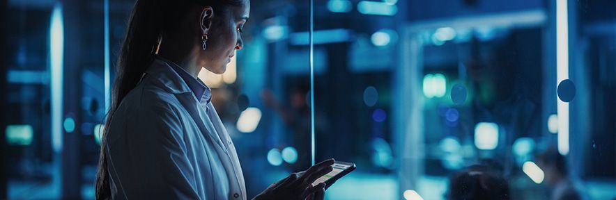 Woman holding a tablet in a blue-lit office. She's wearing a light-colored shirt and looking at the device.
