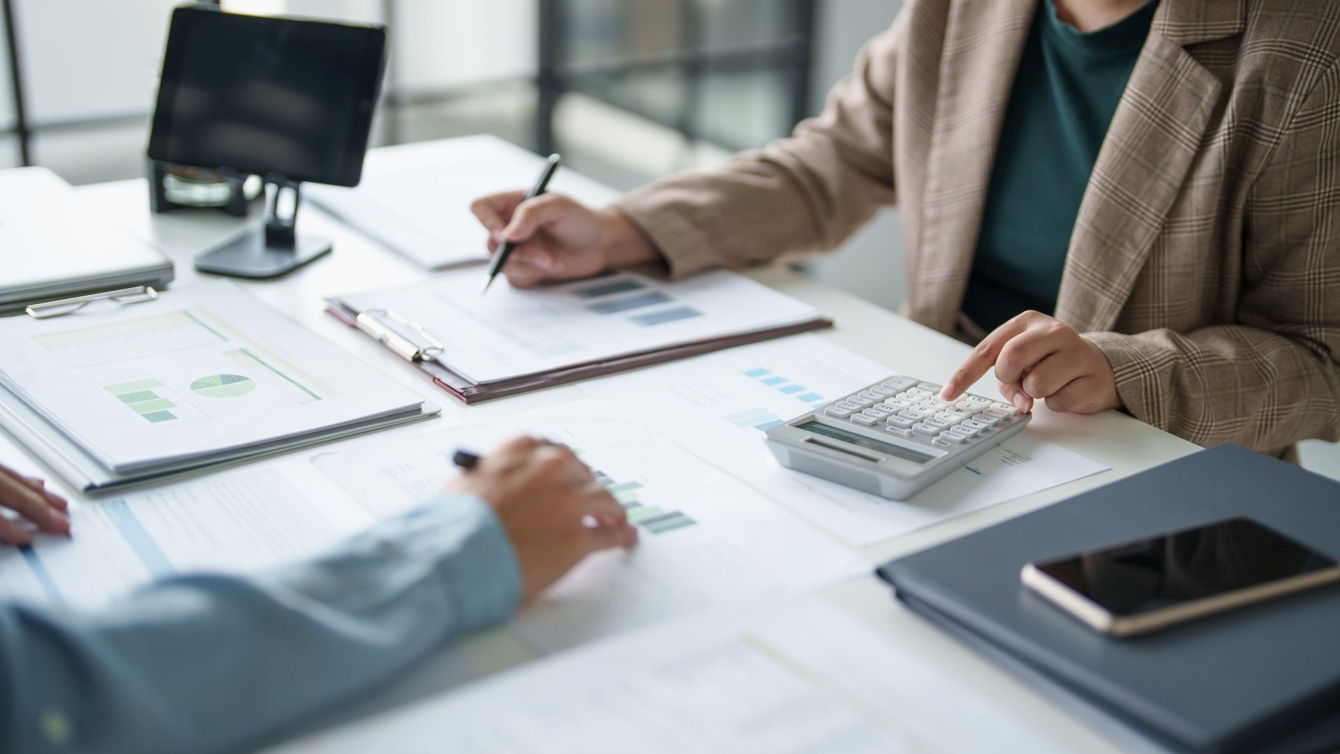 Two people at a desk review financial documents, one using a calculator, in a bright office.