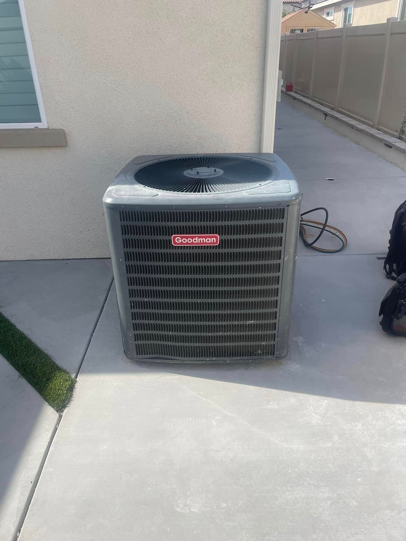 Gray air conditioning unit on a concrete patio next to a house wall and a patch of grass.