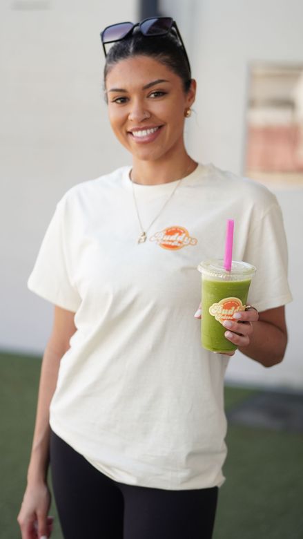 A smiling person wears a white graphic t-shirt and sunglasses, holding a green smoothie with a pink straw.