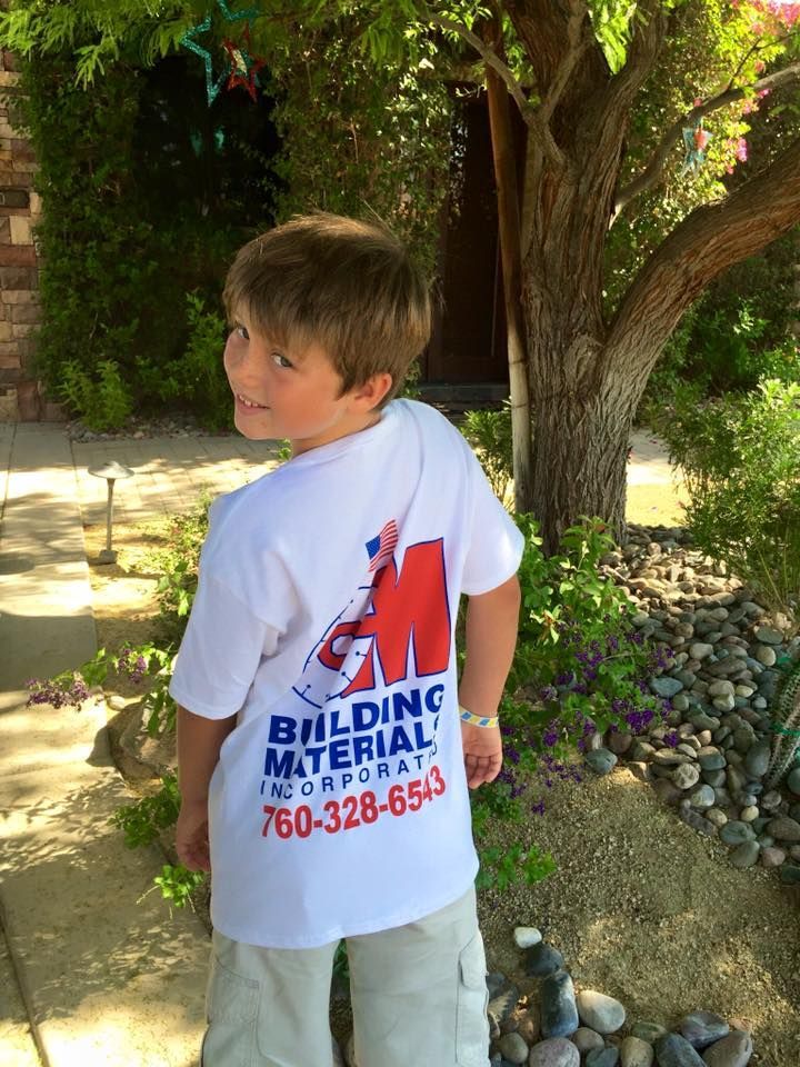 A young boy wearing a building material t-shirt is standing in front of a tree.
