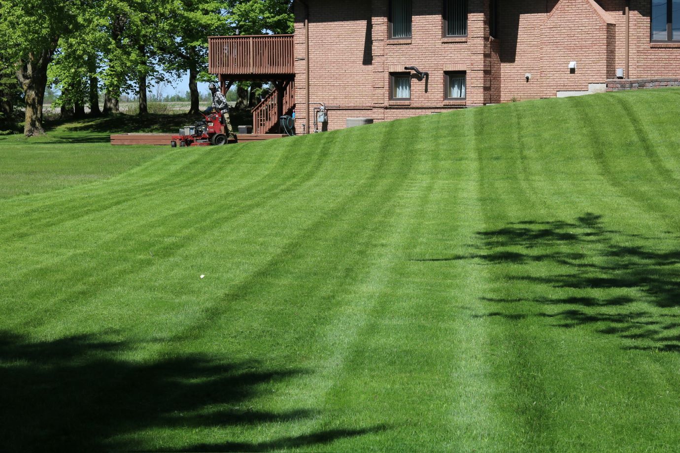 Lawn mower cutting stripes into a grassy hill, with a brick house and deck in the background.