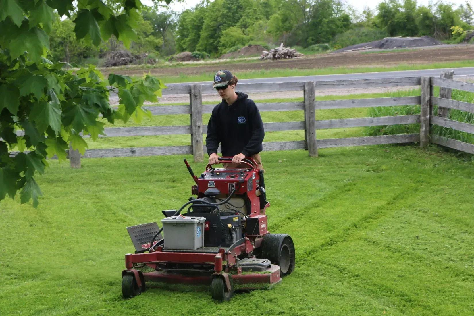 Person mowing a lawn with a red zero-turn mower in a grassy yard, wooden fence in the background.