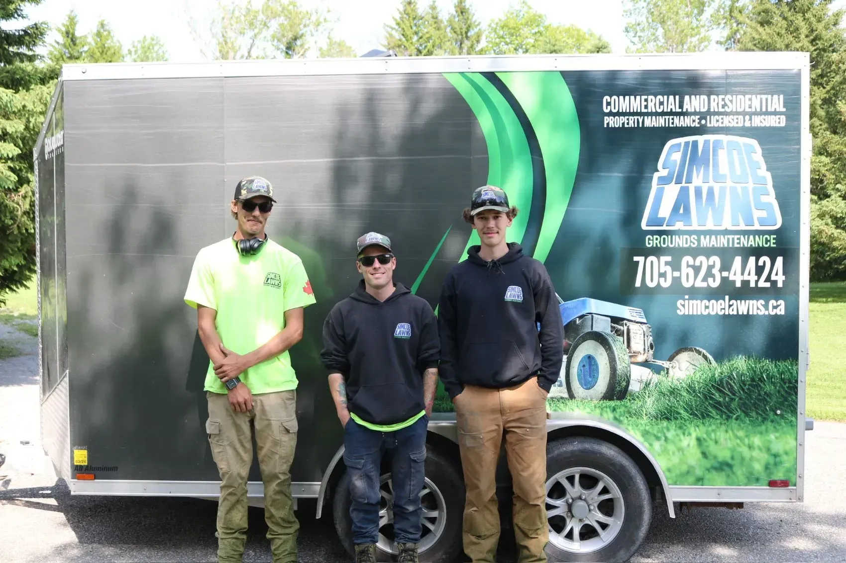 Three people pose in front of a trailer with a lawn care company logo.